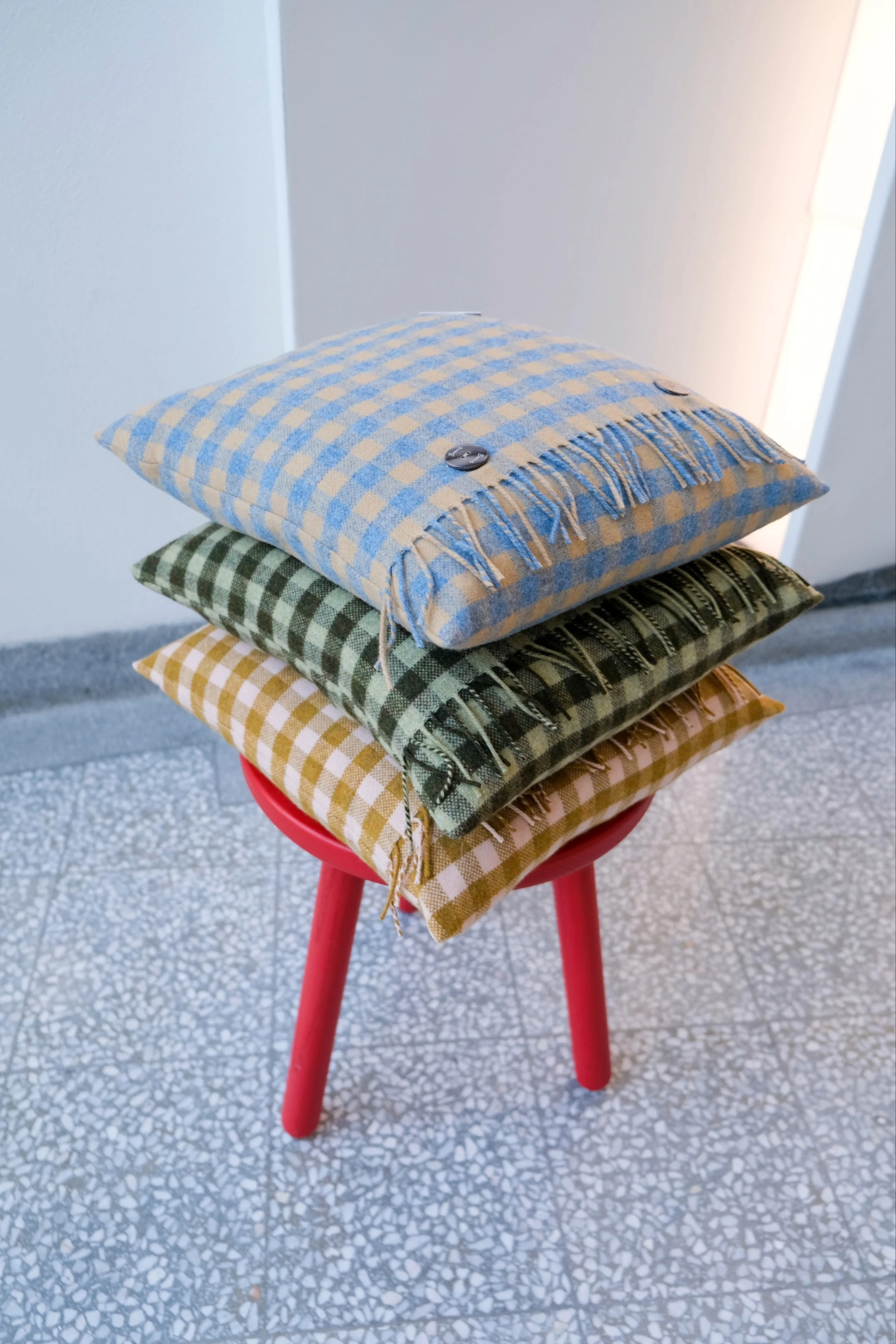 Three folded checkered fabric pillows in yellow, green, and blue, with buttons, stacked on a red stool.