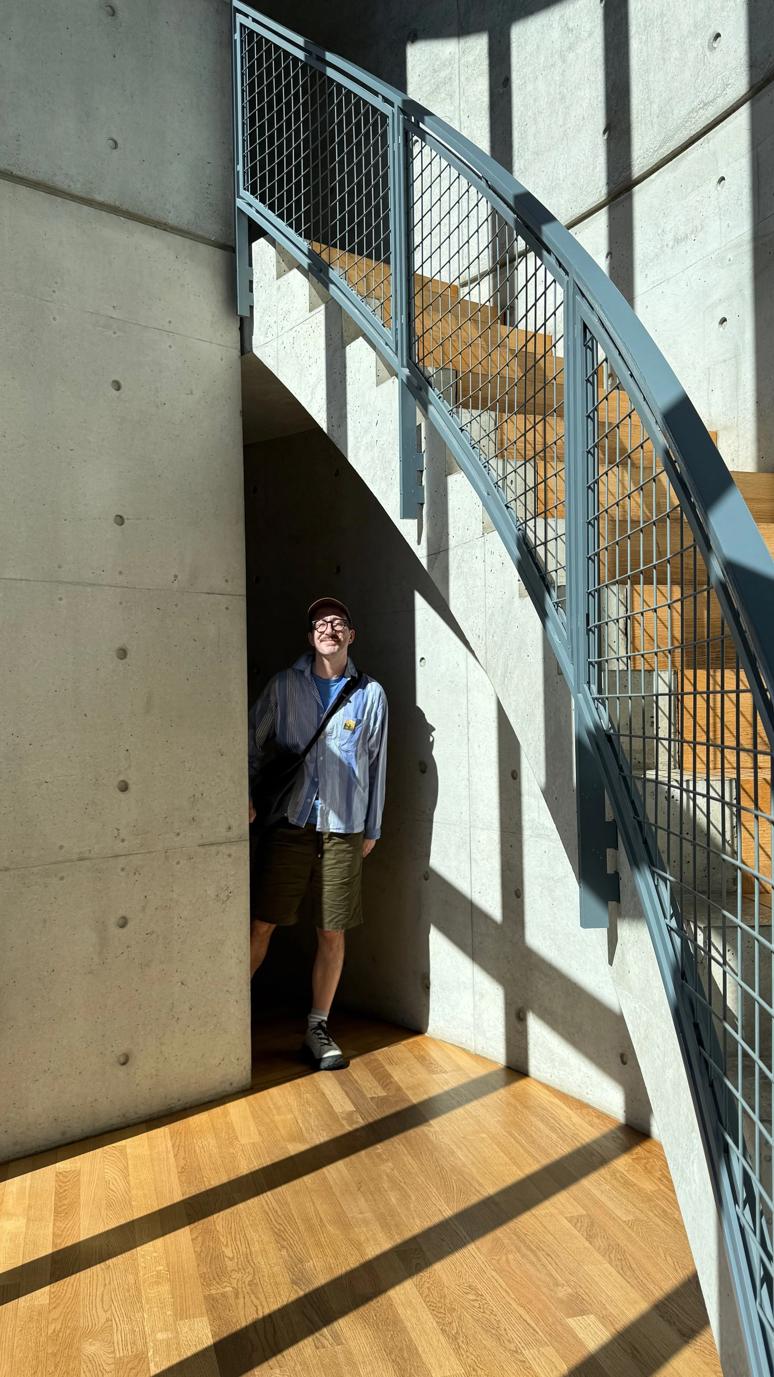A man standing in a shadowed recess of a modern building with concrete walls and wooden flooring, near a curved staircase with metal railings.