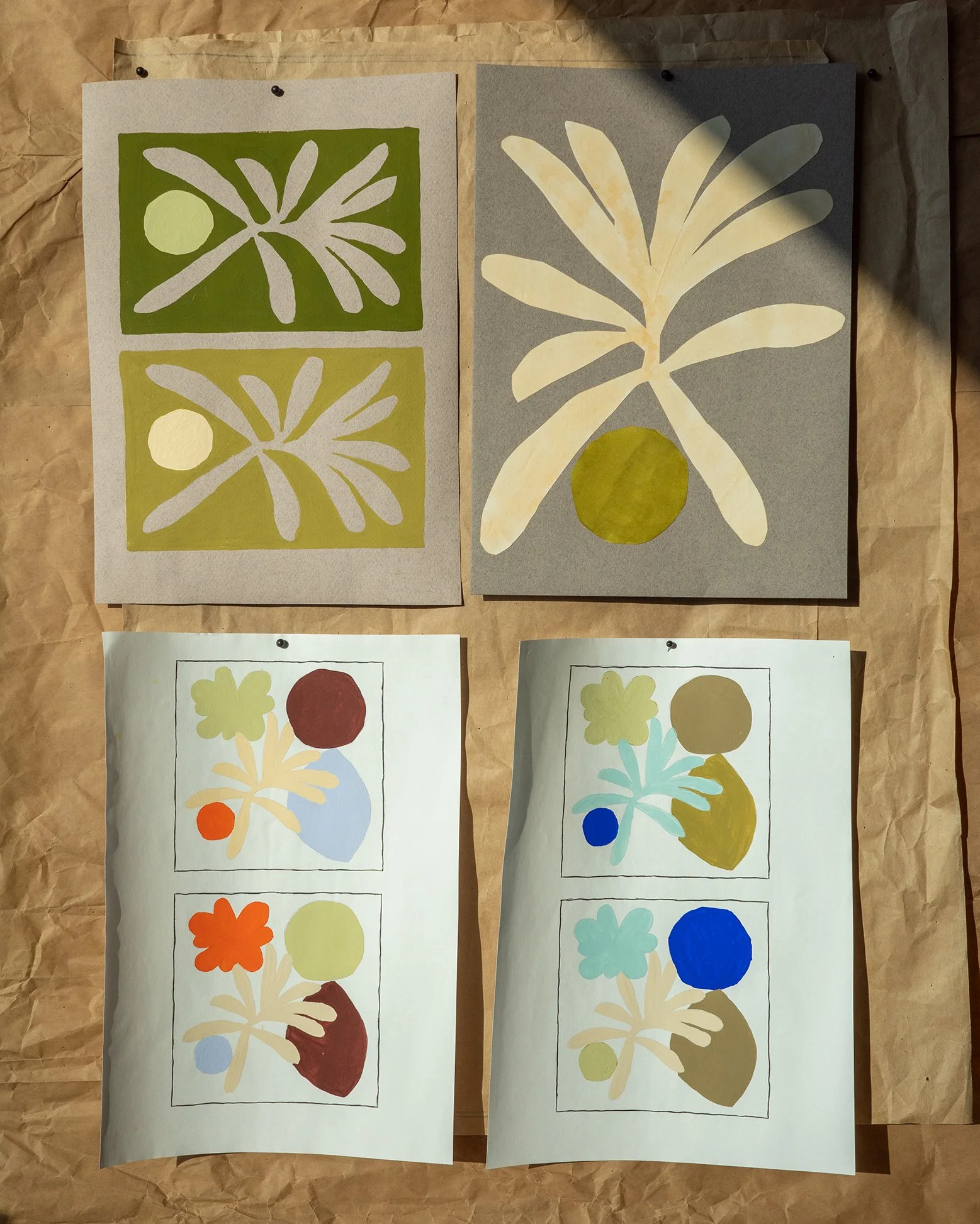 Four colorful nature-themed art prints on paper pinned to a brown sheet with black tacks. The top two prints feature stylized leaves and circles with a background of green and gray, respectively. The bottom prints are features abstract shapes, flowers, and circles in various colors.