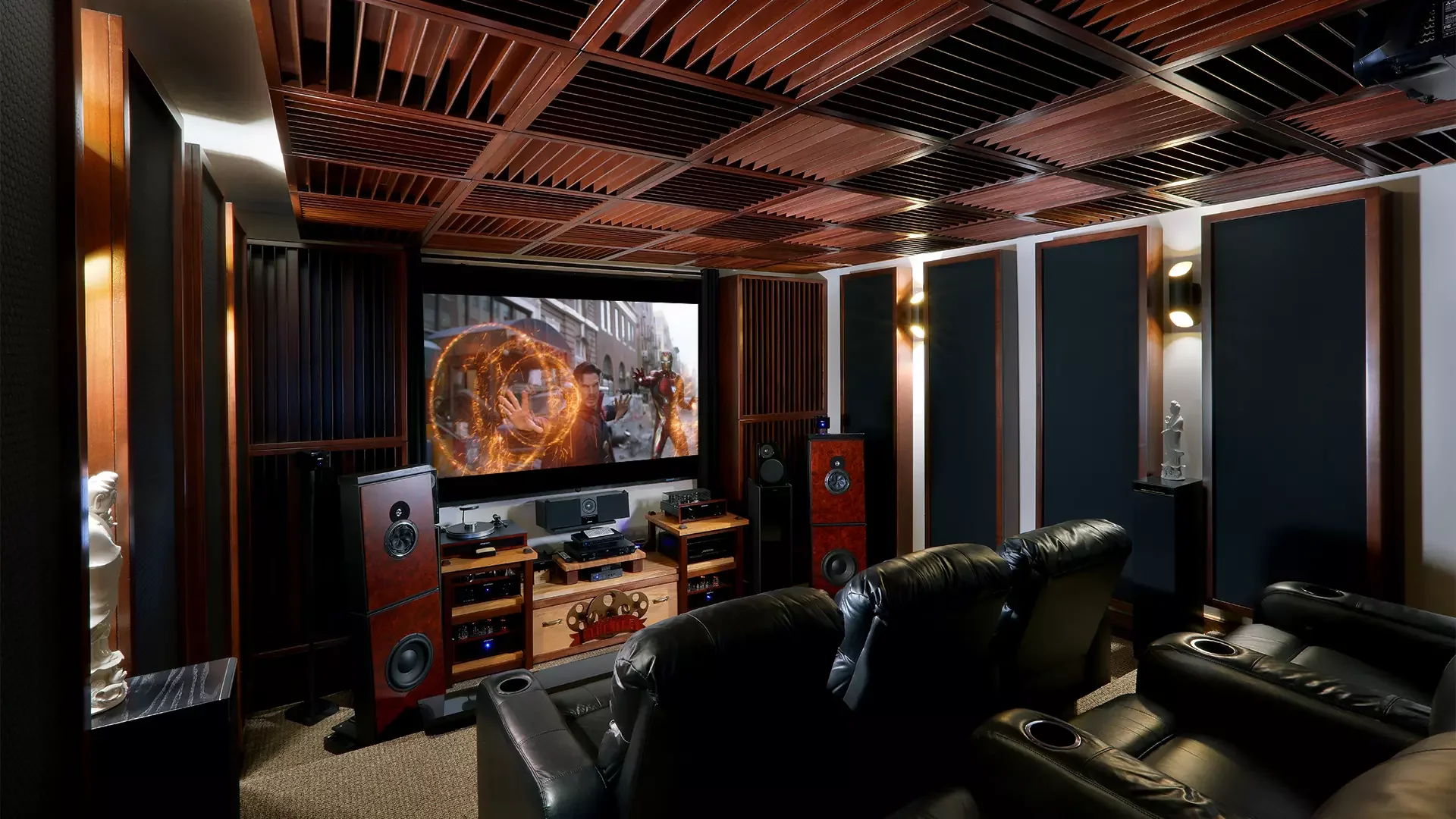 A home theater room with black leather recliner chairs, wooden ceiling panels, and a large flat-screen TV displaying a scene with a person and Iron Man in a city. Surround sound speakers and decorative statues are also visible.
