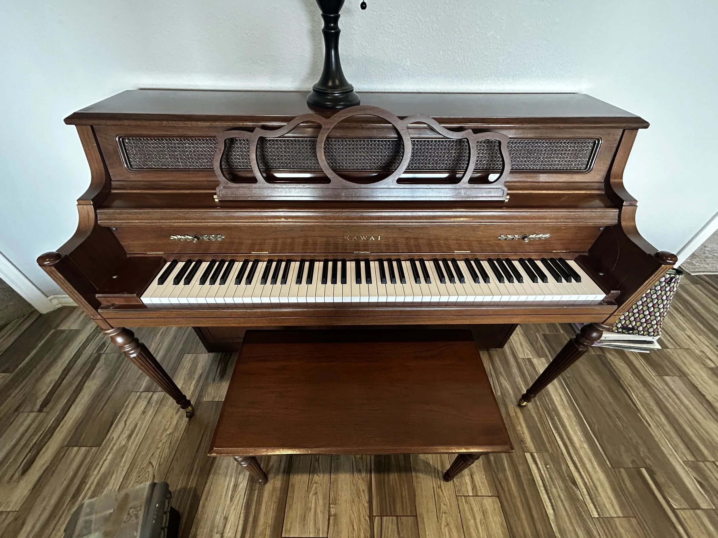 An antique wooden piano with a decorative wooden music stand on top, placed against a light-colored wall, with a hardwood floor beneath.