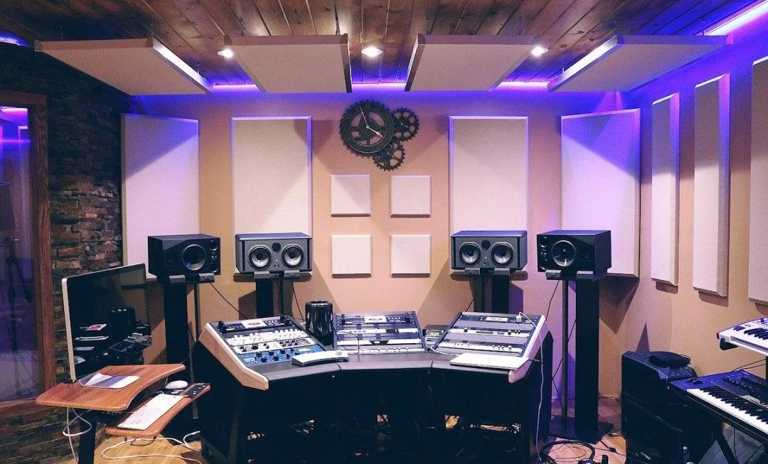 Music recording studio with mixing console, speakers, and electronic equipment, illuminated with purple lighting.
