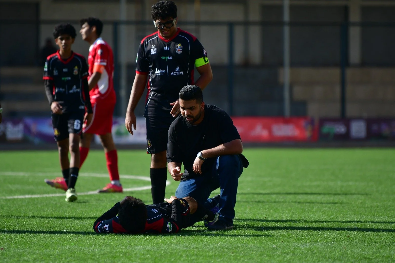 A soccer player is lying on the field, injured, as a man checks on him. Two other players and a referee observe from a distance on the field.