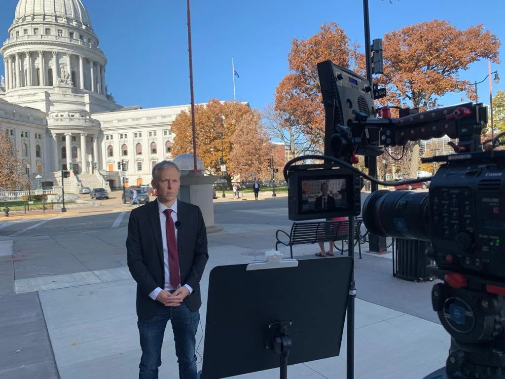 Ein Mann in Anzug steht vor der Kamera, während im Hintergrund das State Capitol in Madison, Wisconsin zu sehen ist, umgeben von Herbstbäumen.