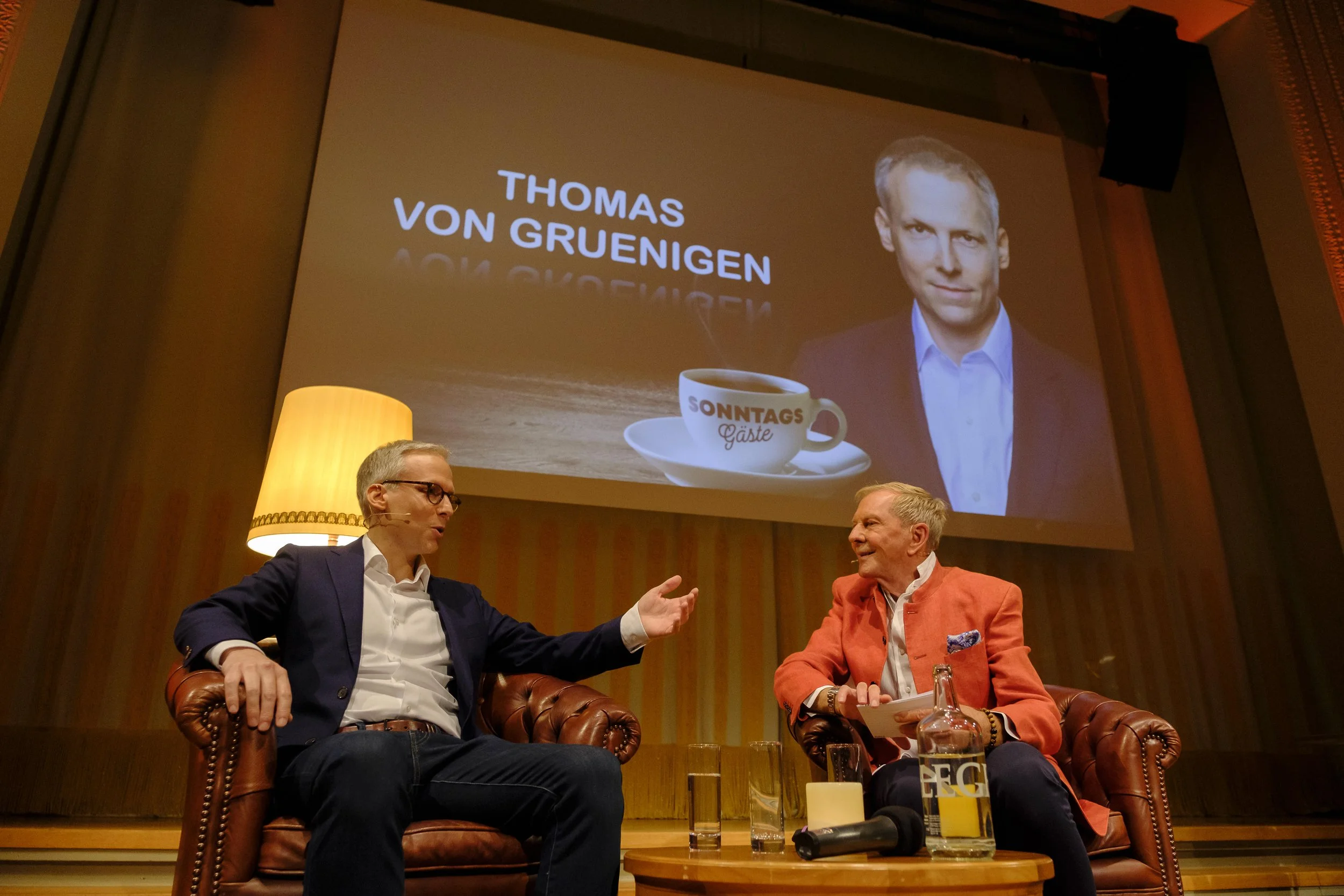 Zwei Männer führen ein Gespräch auf einer Bühne, im Hintergrund ein projiziertes Bild mit Namen 'Thomas von Gruenigen', einer Kaffeetasse und einem Gesicht. Die Szene spielt in einem stilvollen Raum mit warmem Licht, einer Stehlampe und einem kleinen Tisch mit Getränken.
