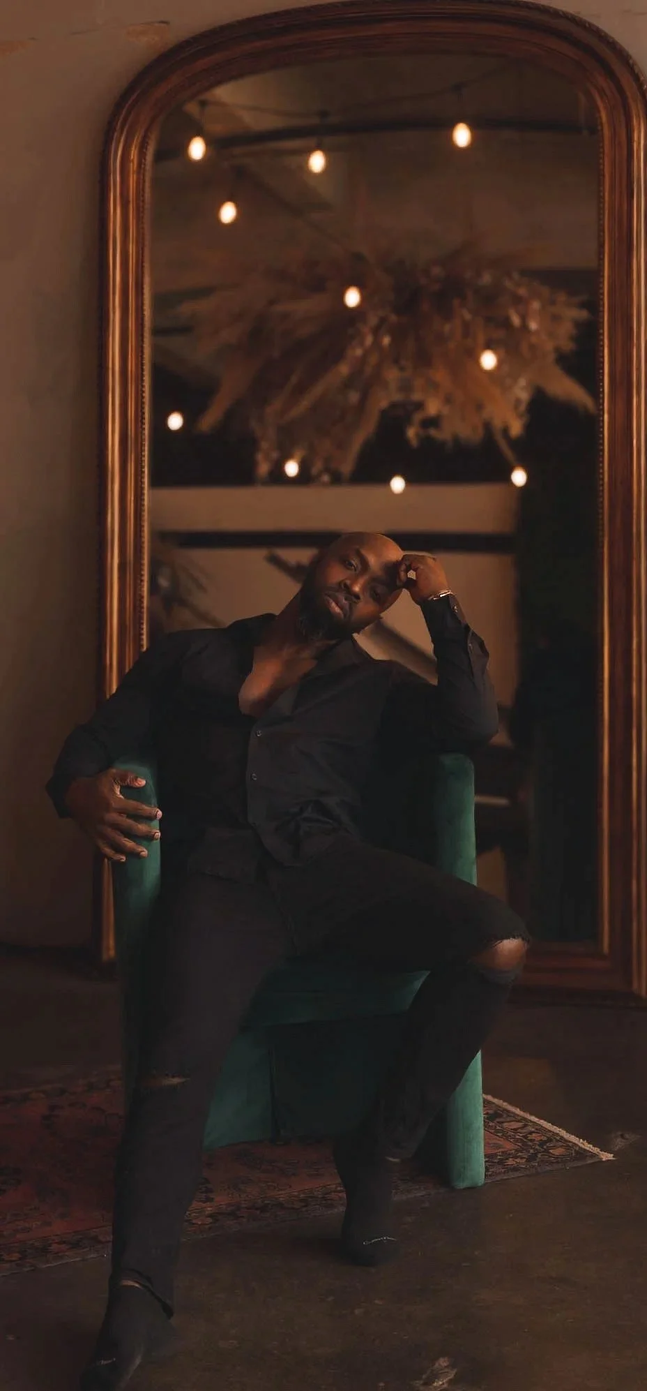 A man in black clothing sitting on a teal chair, leaning his head on his hand, in front of a large, ornate gold mirror with a reflection of a chandelier with multiple bulbs. Male Boudoir