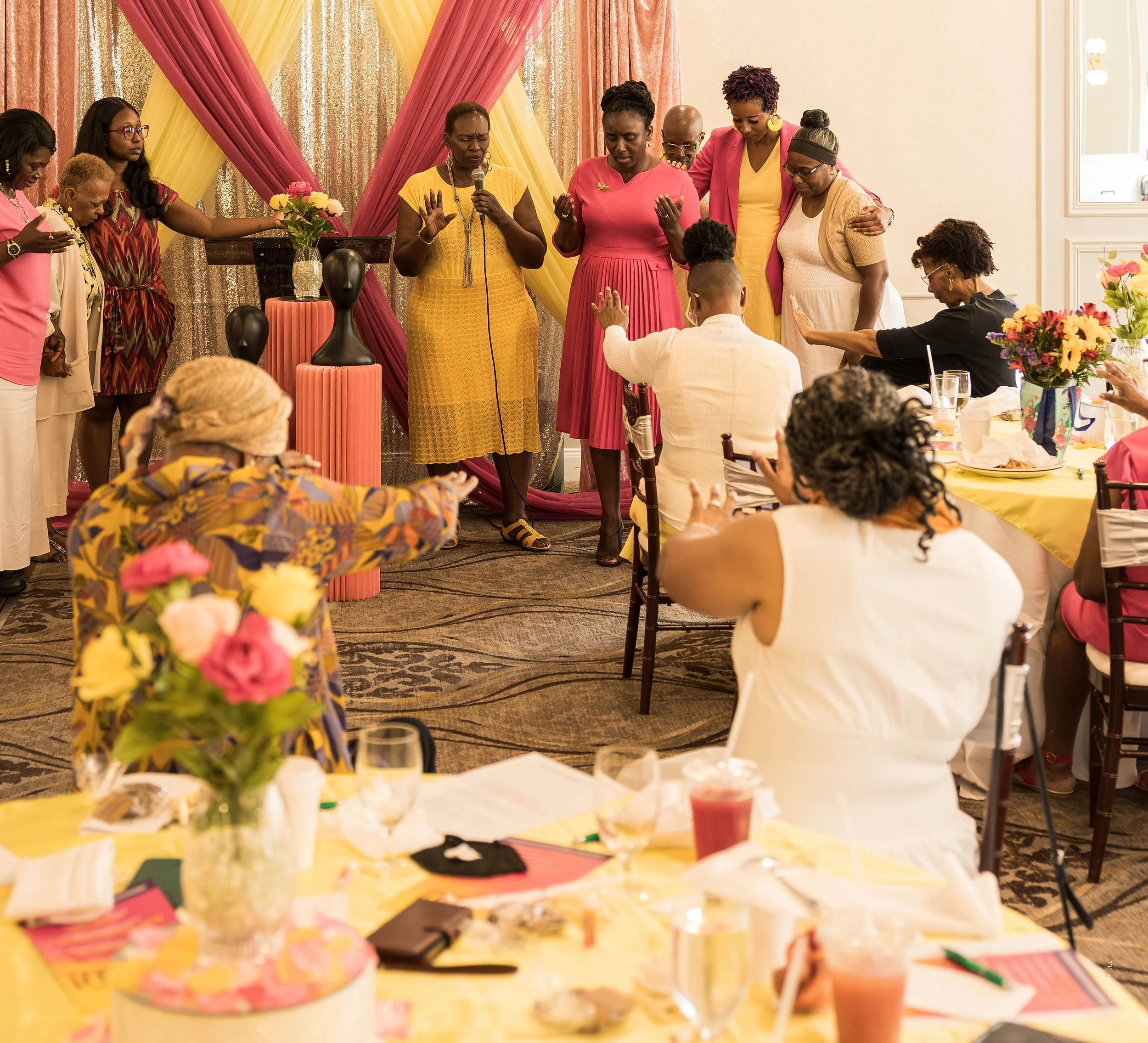 A group of women praying with heads bowed and eyes closed at a celebration or gathering, with colorful decorations and flowers in the background. Ministry Event