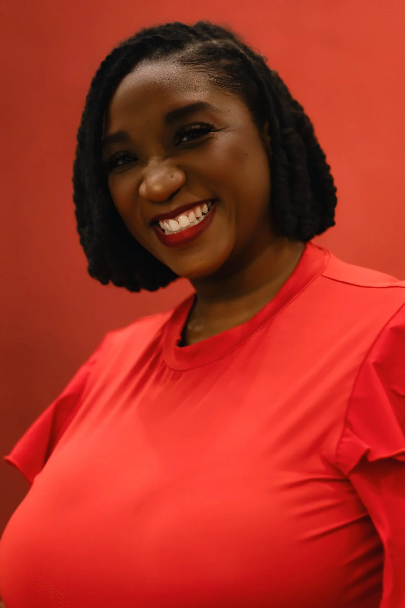 A smiling woman with dark, shoulder-length twisting hair wearing a red top against a matching red background. Portrait