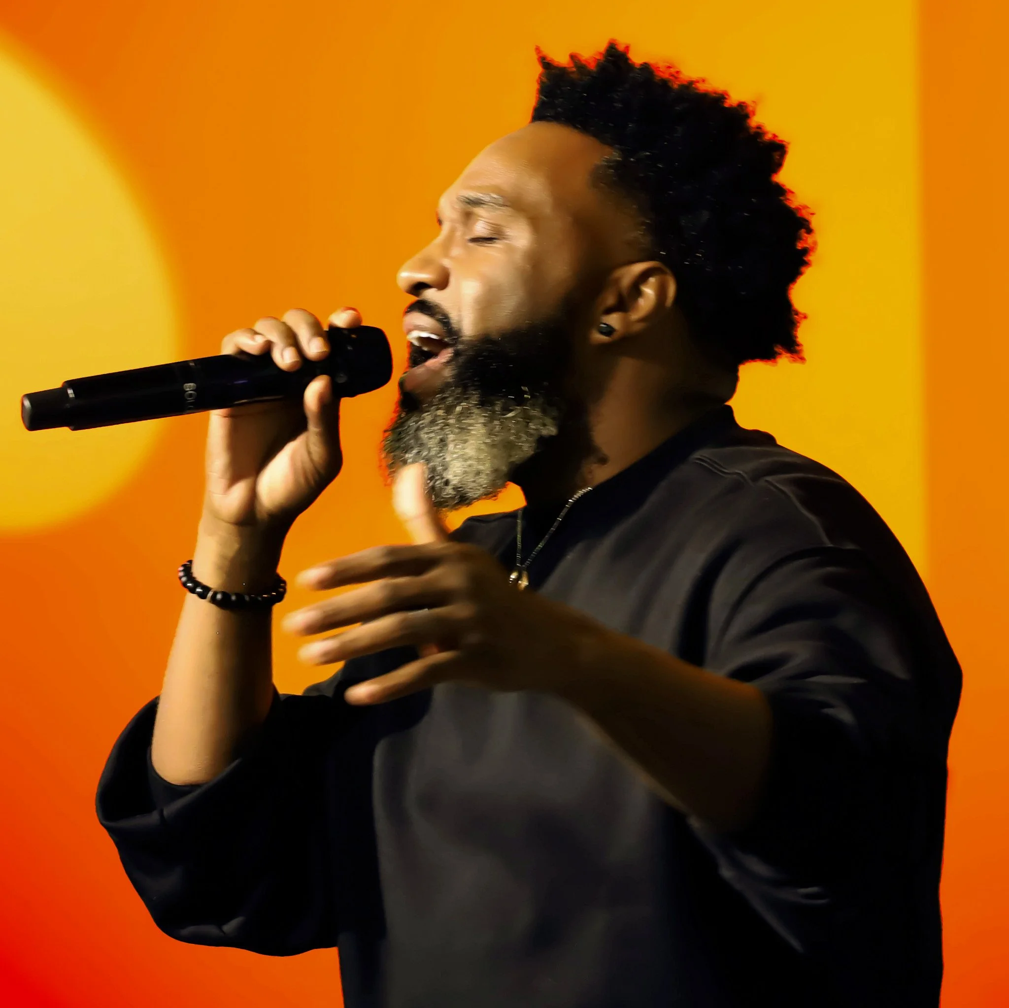 A man with a beard singing into a microphone against an orange background. Event