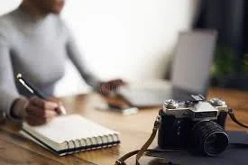 Person writing in a notebook at a desk with a camera in the foreground and a laptop in the background