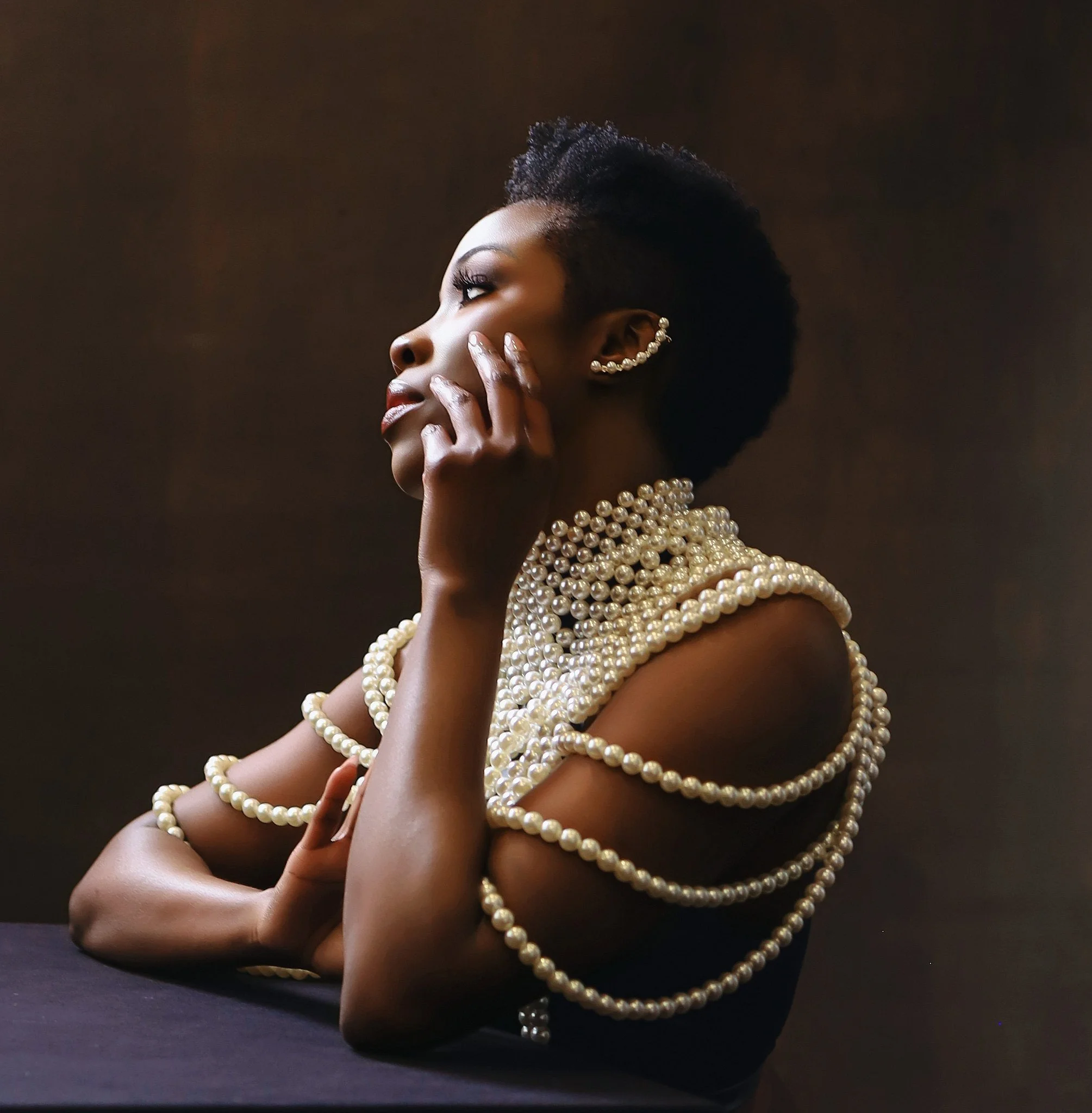 A woman with short natural hair, wearing pearl earrings and multiple strands of pearl necklaces, posed with her hand on her face against a dark background. Portrait