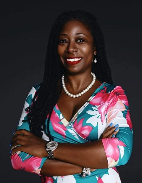 A woman with dark skin and long braided hair smiling confidently, wearing a colorful floral dress, pearl necklace, earrings, and a wristwatch, against a dark background. Headshot