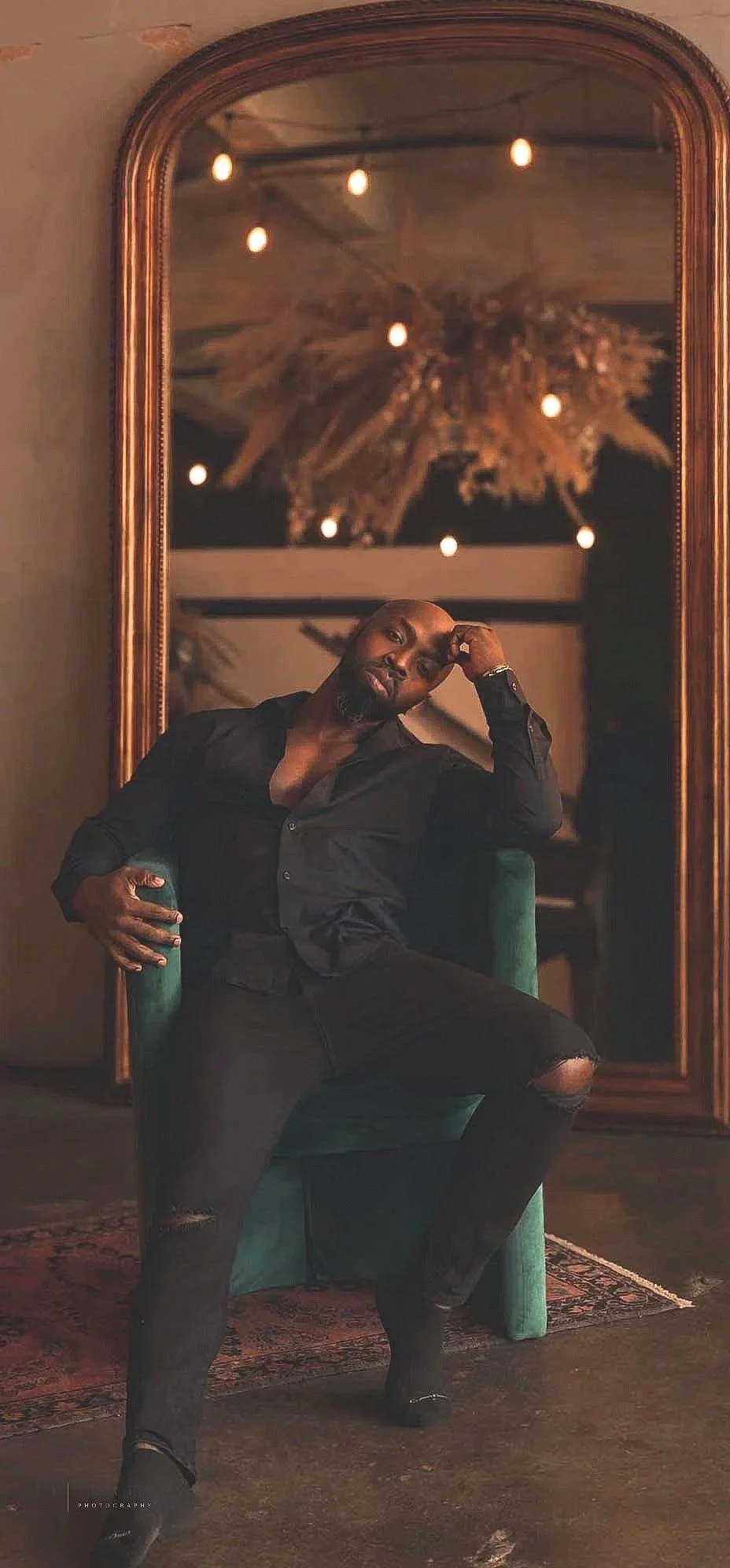 A man sitting on a green chair in front of a large, ornate mirror, with a chandelier and warm lighting reflected in the mirror.
