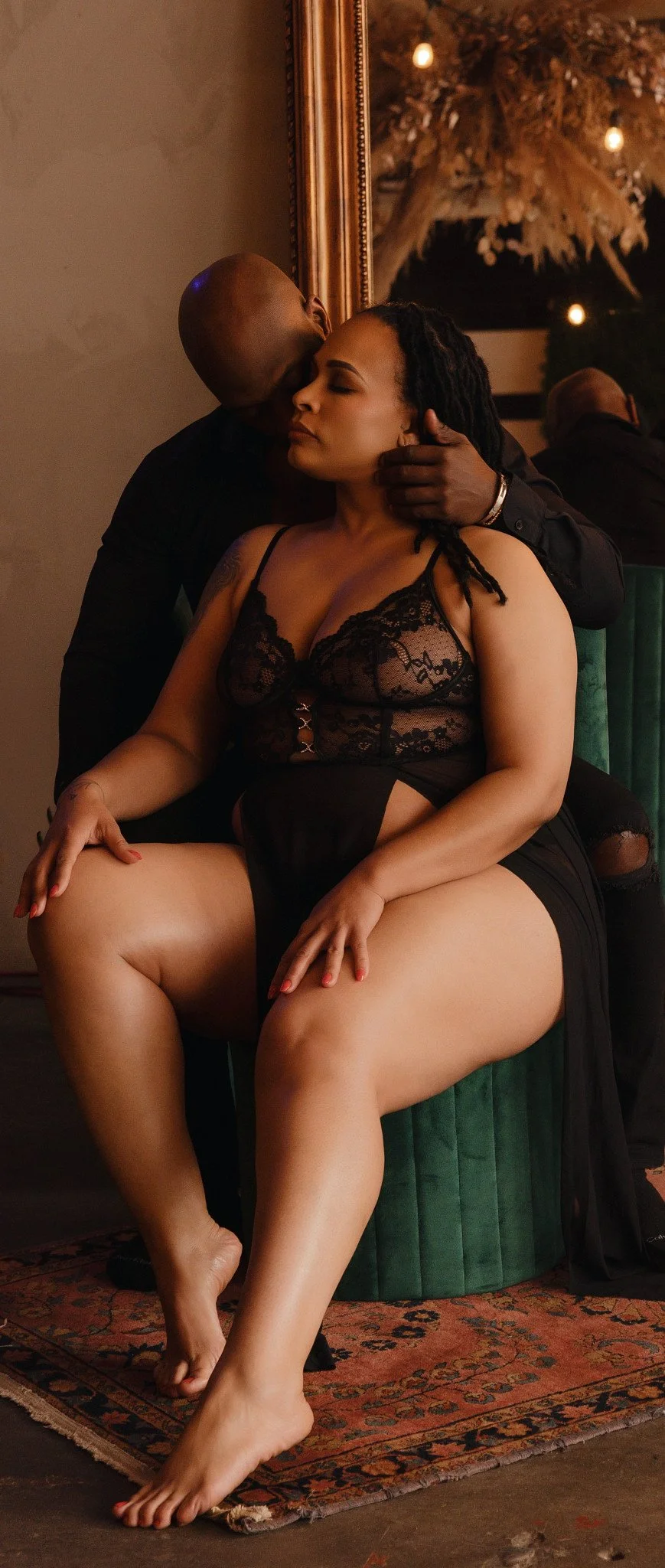 A woman in black lace lingerie sitting on a green velvet chair, while a man in black clothing leans in to kiss her cheek in a dimly lit room with warm lighting and a large mirror in the background. Couple Boudoir
