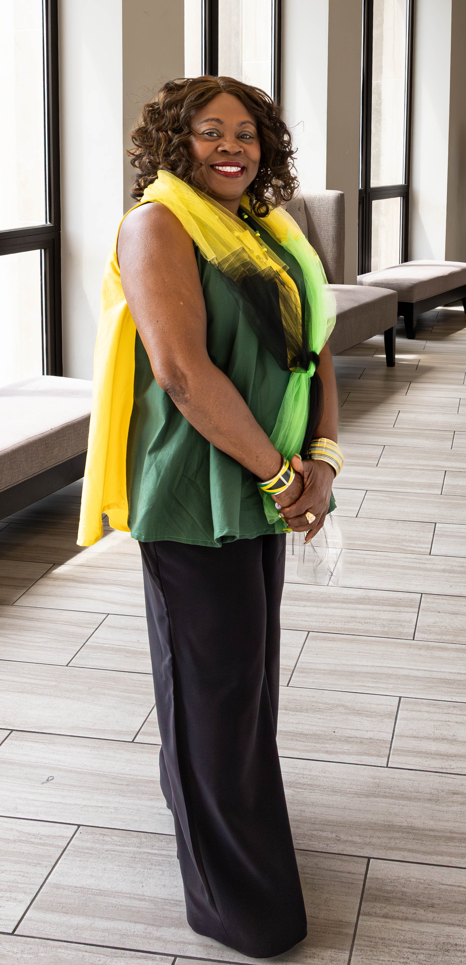 A smiling woman with curly hair wearing a green shirt and black pants, accessorized with yellow and green jewelry, standing inside a modern building with large windows and light-colored tiled floors.