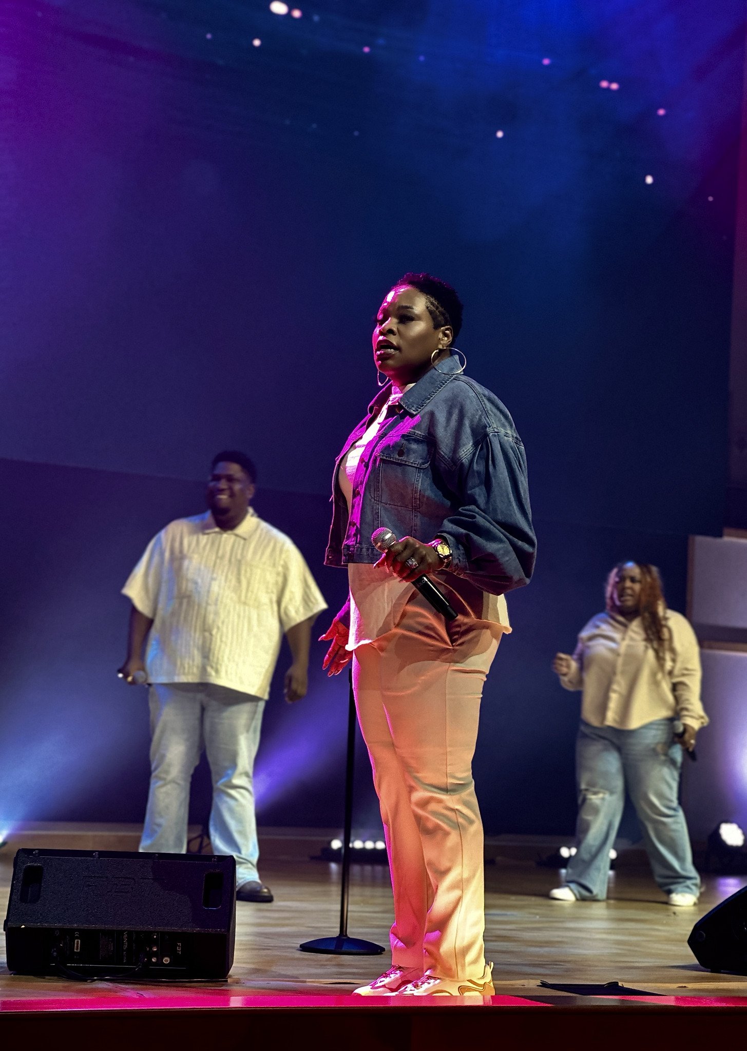 A woman in beige pants and a denim jacket holding a microphone and singing on stage, with three backup singers behind her, under colorful stage lighting. Event