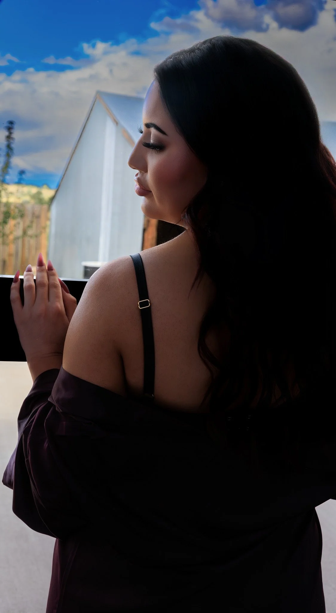 A woman with long dark hair and makeup, wearing a dark off-shoulder top and a thin strap undergarment, with her hands clasped together, looking down and to the side, by a window with a view of a blue sky with clouds and a building outside.
