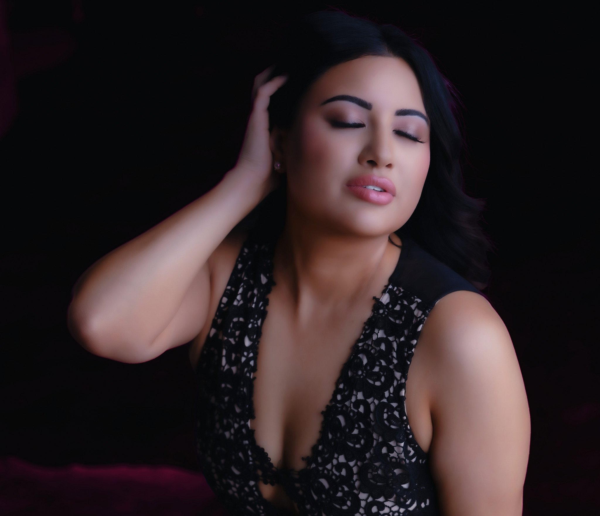 Close-up of a woman with long dark hair, closed eyes, wearing subtle makeup, wearing a black dress with white floral pattern, and resting her hand on her head. Portrait