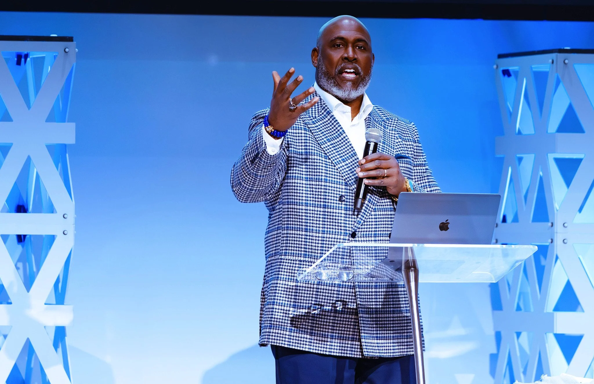A bald man with a gray beard wearing a checkered jacket and white shirt is speaking into a microphone while standing behind a clear podium at a conference or event.