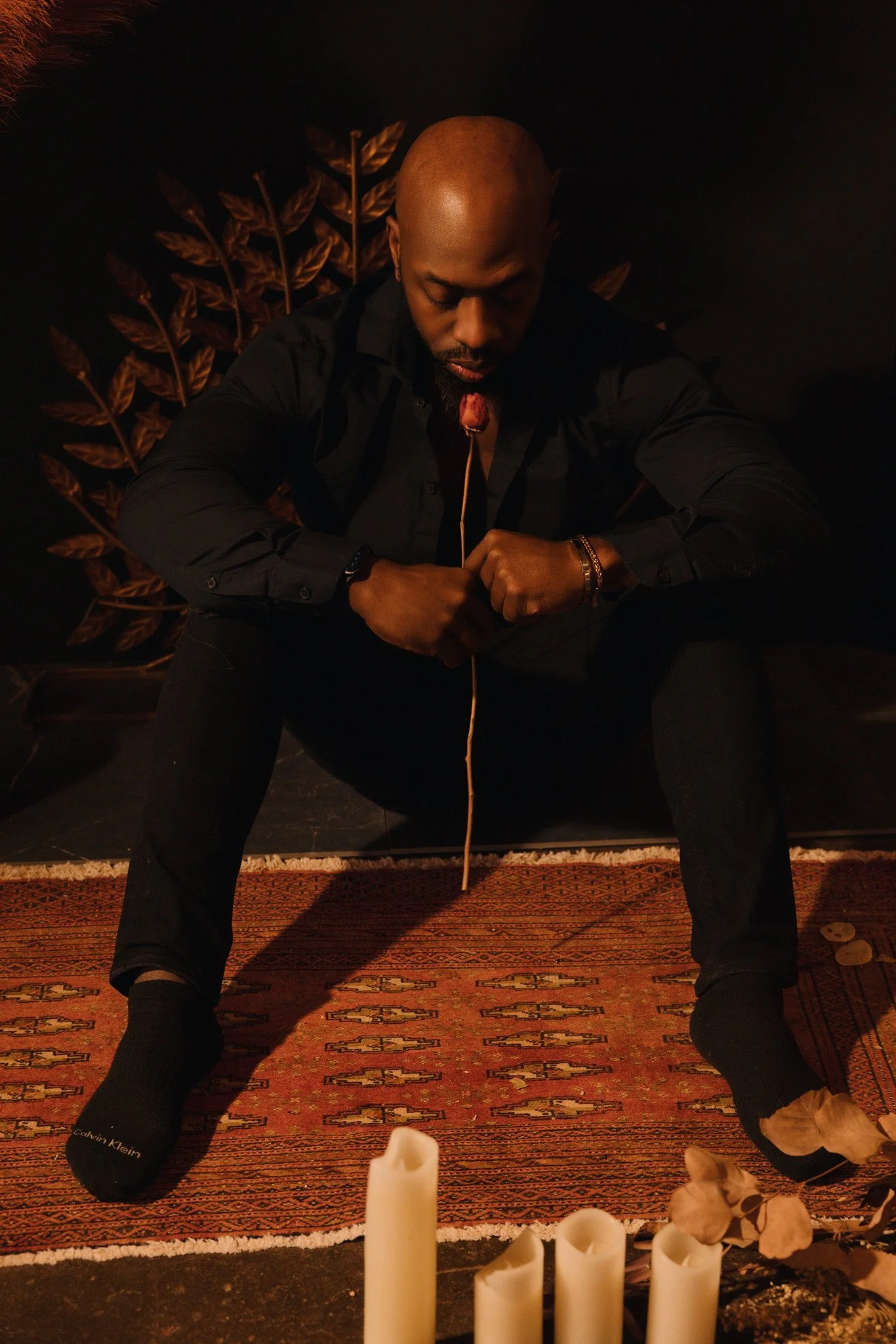 A man sitting on a rug with candles and flowers in front of him, wearing black Calvin Klein socks, with his head bowed and eyes closed, in a dimly lit room with dark background and decorative wall art.