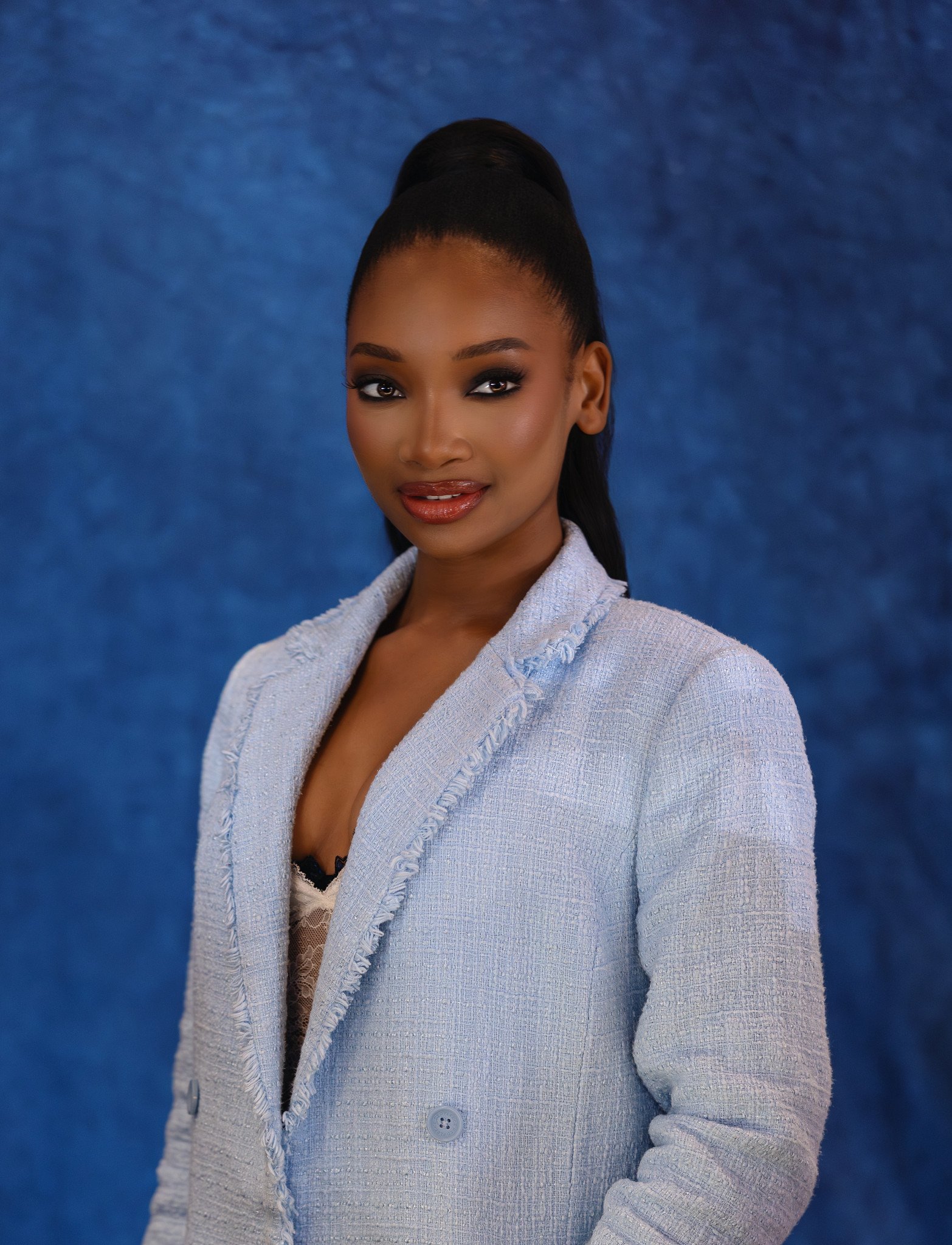 Close-up of a woman with dark hair styled in a high ponytail, wearing makeup, a white textured blazer, and a black top with lace, standing against a blue background.