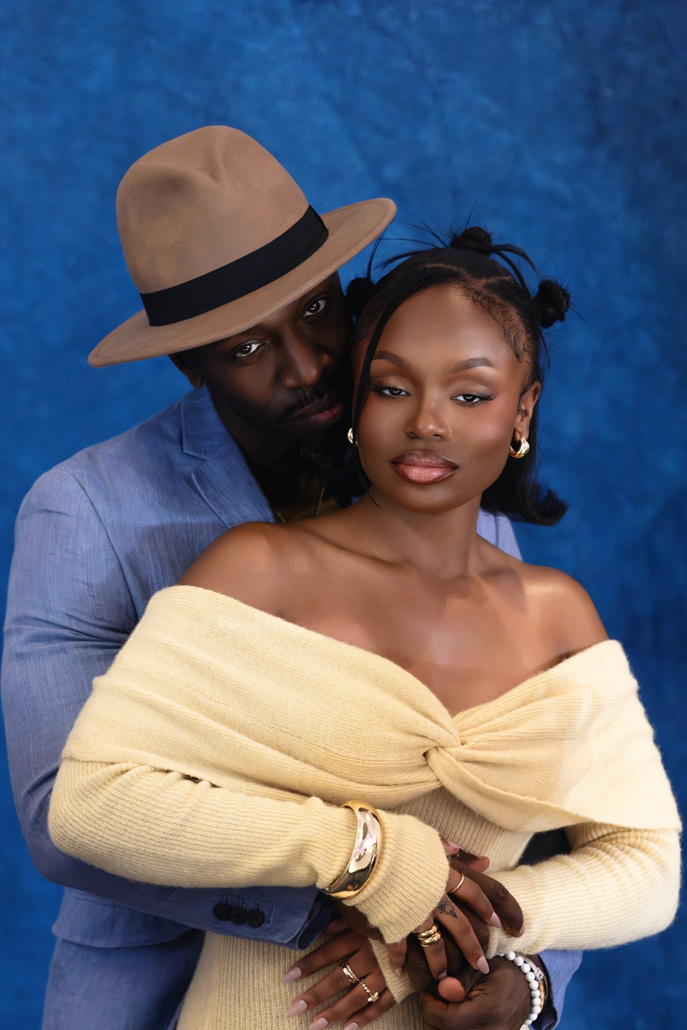 A couple embracing, the man wearing a beige fedora hat and a light blue suit, and the woman wearing a cream off-shoulder sweater with jewelry, against a blue background. Family Portrait