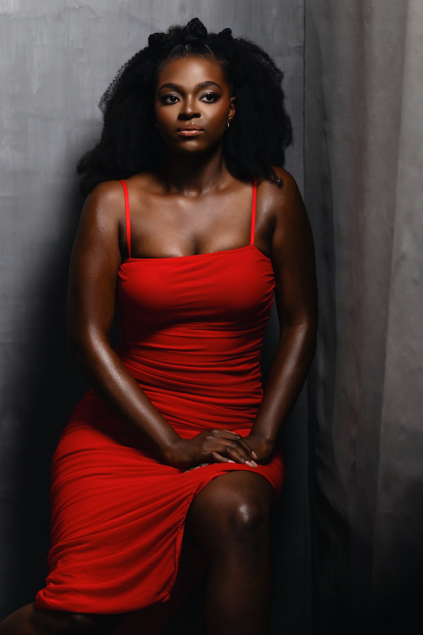 A woman with dark skin and curly black hair styled in an afro, wearing a red dress with thin straps, sitting against a gray textured background. Portrait