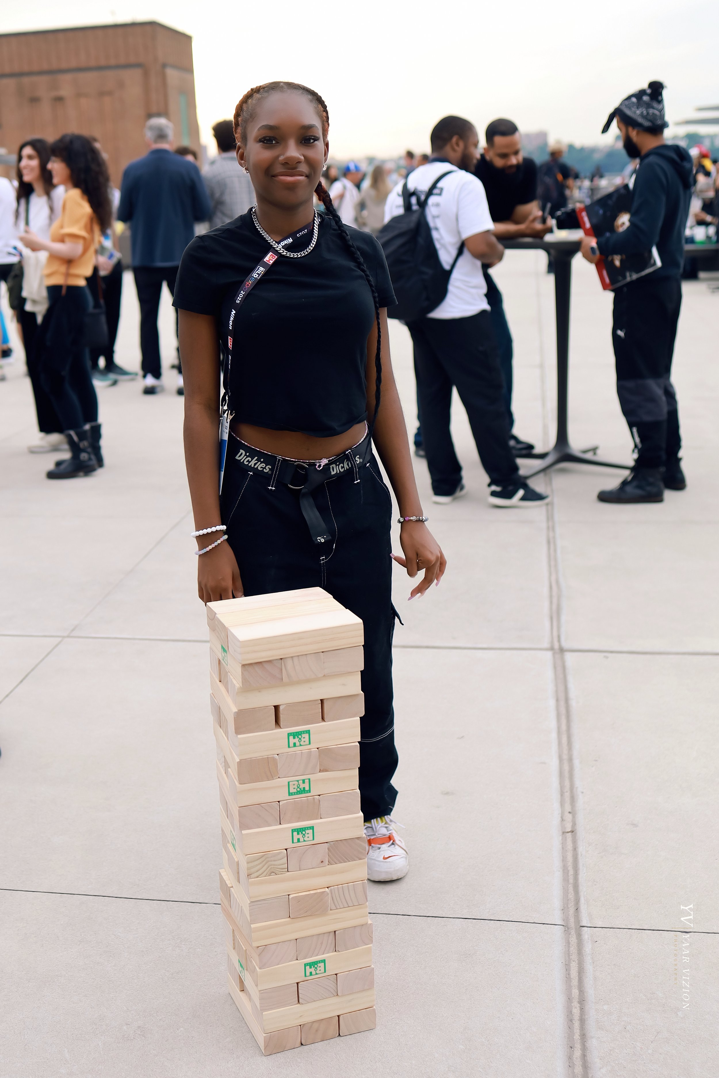 A young woman standing next to a stack of large wooden Jenga blocks outdoors during daytime. The woman has braided hair, is wearing a black crop top, dark pants, and white sneakers. People are gathered in the background, some engaged in conversations