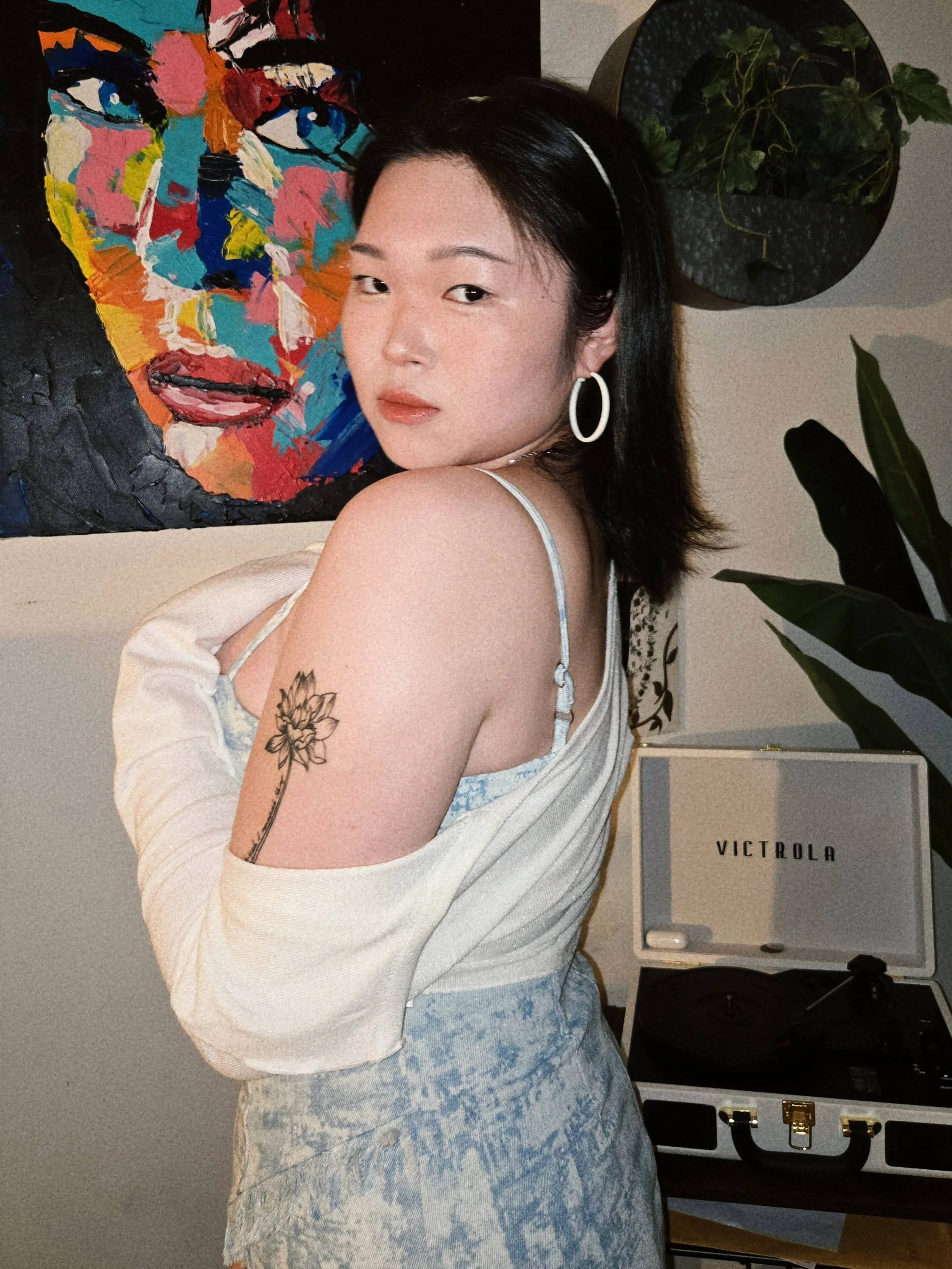 A woman with black hair and a white headband, wearing a light-colored off-shoulder top and light blue jeans, standing in front of a colorful abstract portrait painting on the wall. There are plants and a white Vicrola stereo on a shelf in the background.