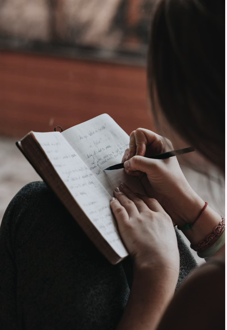 Person writing in a notebook with a pen, seated outdoors with a brick surface in the background.