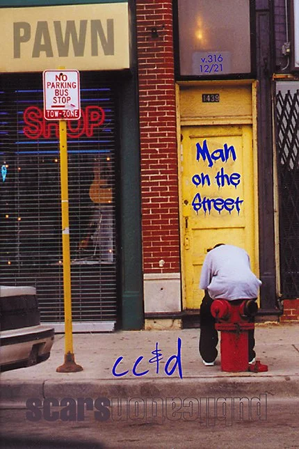 A man in a white hoodie kneeling on the sidewalk at the corner of a yellow building with a blue street art message 'Man on the street'. There is a red fire hydrant and a parking meter nearby. The scene includes a pawn shop with a sign saying 'PAWN' and a parking restriction sign. A no parking sign and a stop sign are also visible.