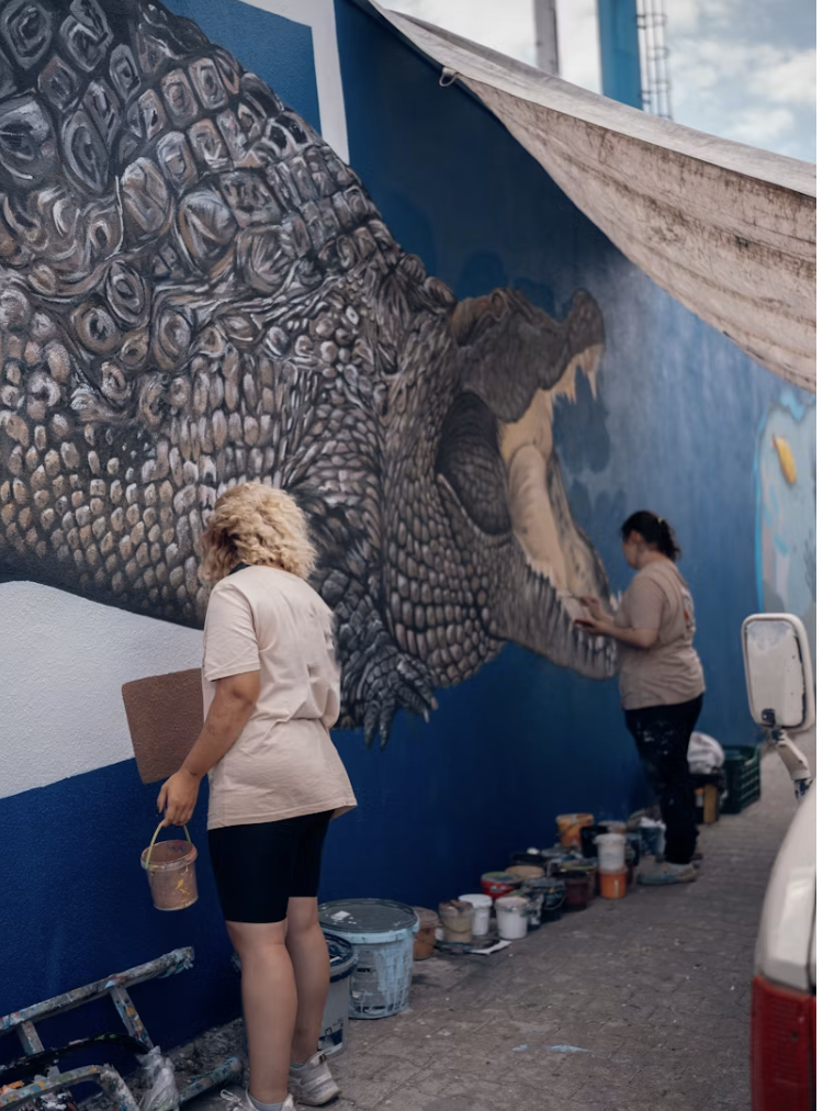 Two artists painting a mural of a dinosaur on a blue wall.