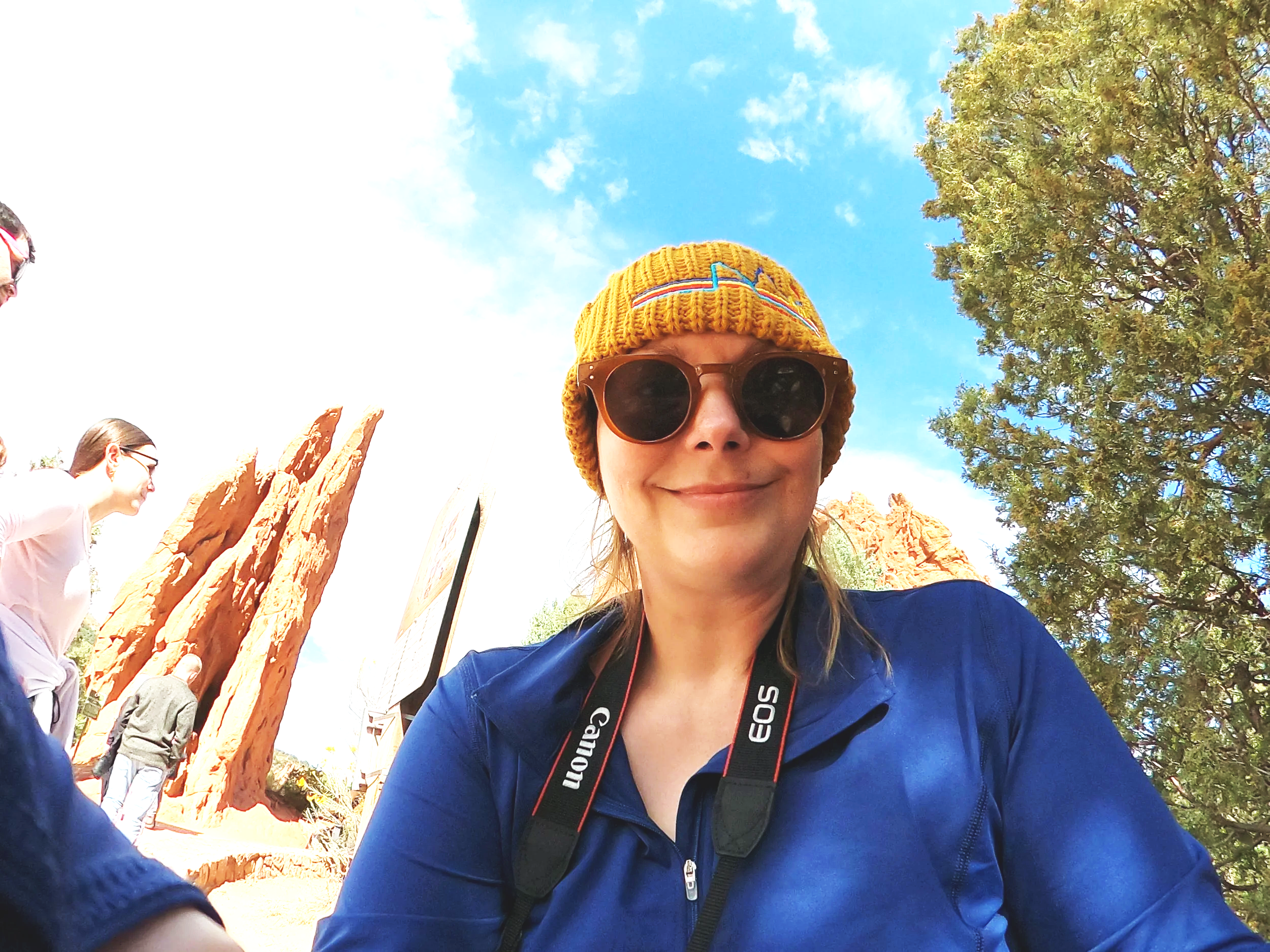 A woman wearing sunglasses, a yellow knit hat, and a blue jacket smiling at the camera outdoors during the day. In the background, there are other people, orange red rock formations, a green tree, and a partly cloudy sky of Colorado Springs, Colorado
