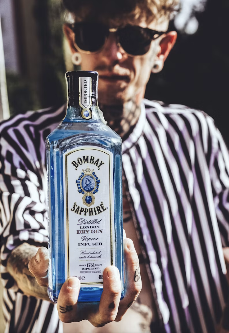 A person with glasses and tattoos holding a bottle of Bombay Sapphire gin in front of them, with a blurred outdoor background.