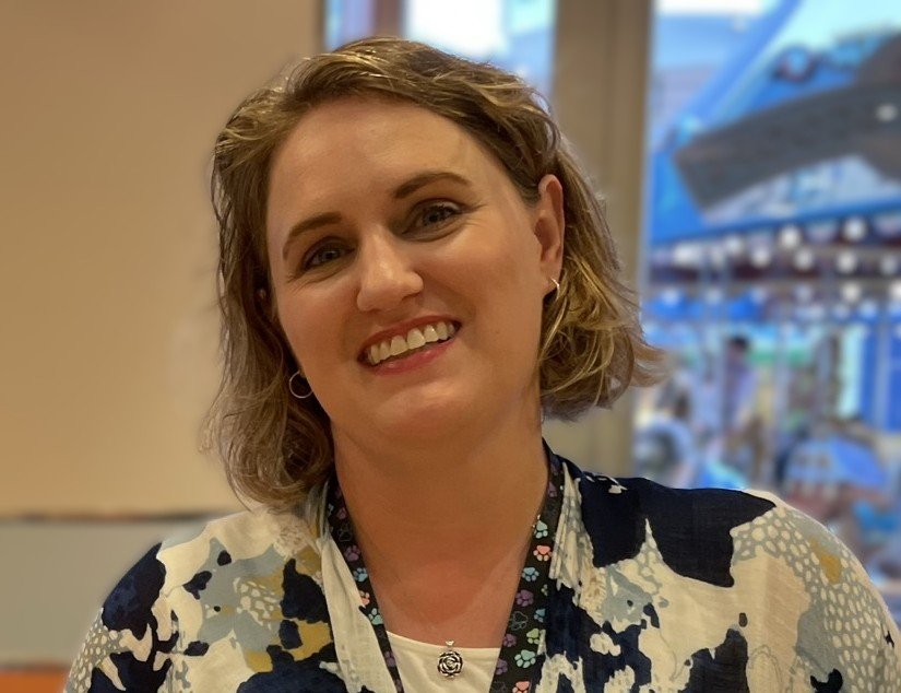 A smiling woman with short, wavy blonde hair, wearing a black and white patterned top, earrings, and a lanyard, in an indoor setting with a window showing an outdoor cafe scene.