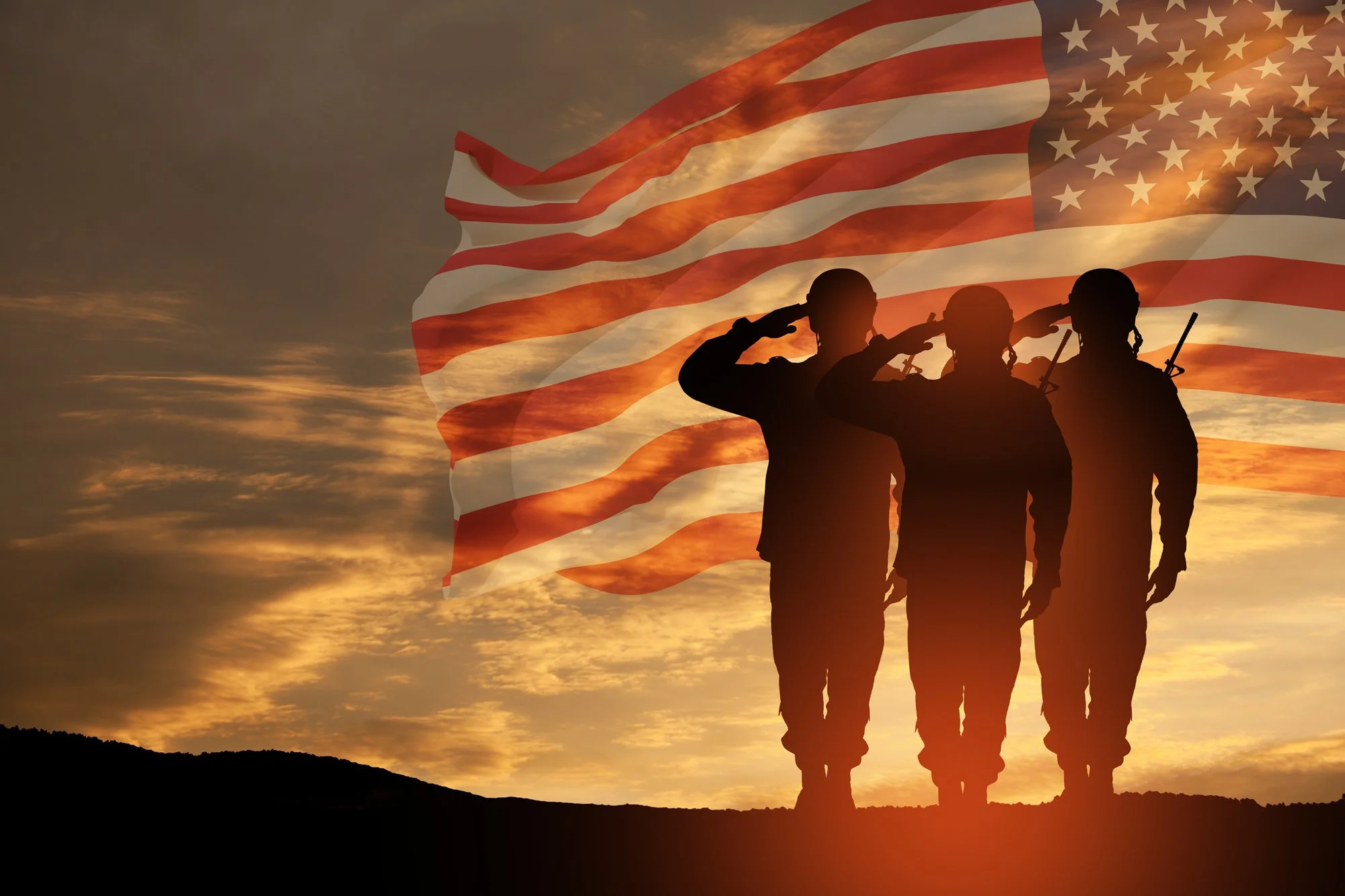 Three soldiers saluting during sunset with an American flag in the background.