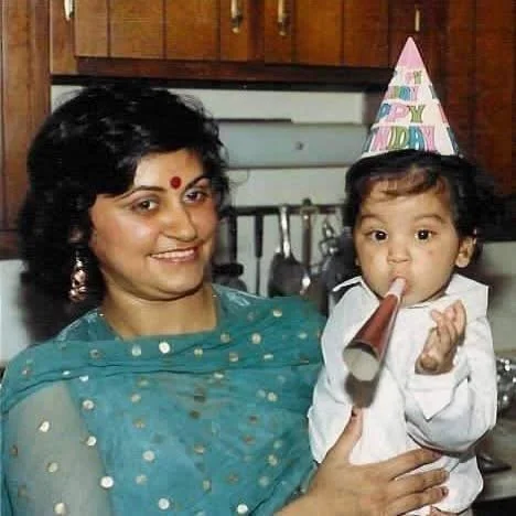 A woman holding a young Rohit Malhotra at a birthday celebration, with the child wearing a birthday hat and blowing a party blower.