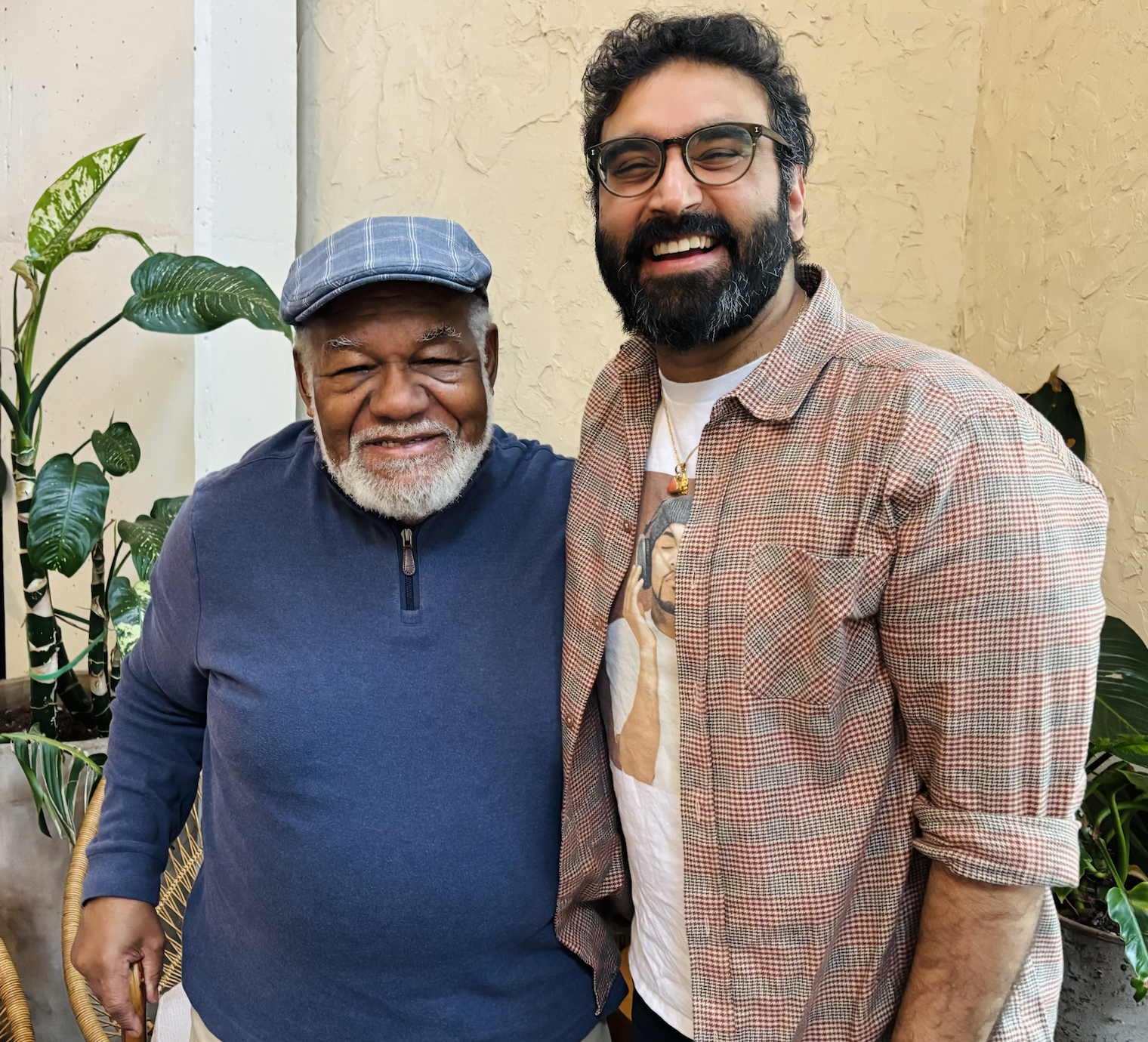 Two men smiling and posing together indoors. One is an older man with gray hair and beard, wearing a blue zip-up sweater and a blue plaid cap. The other is Rohit Malhotra.