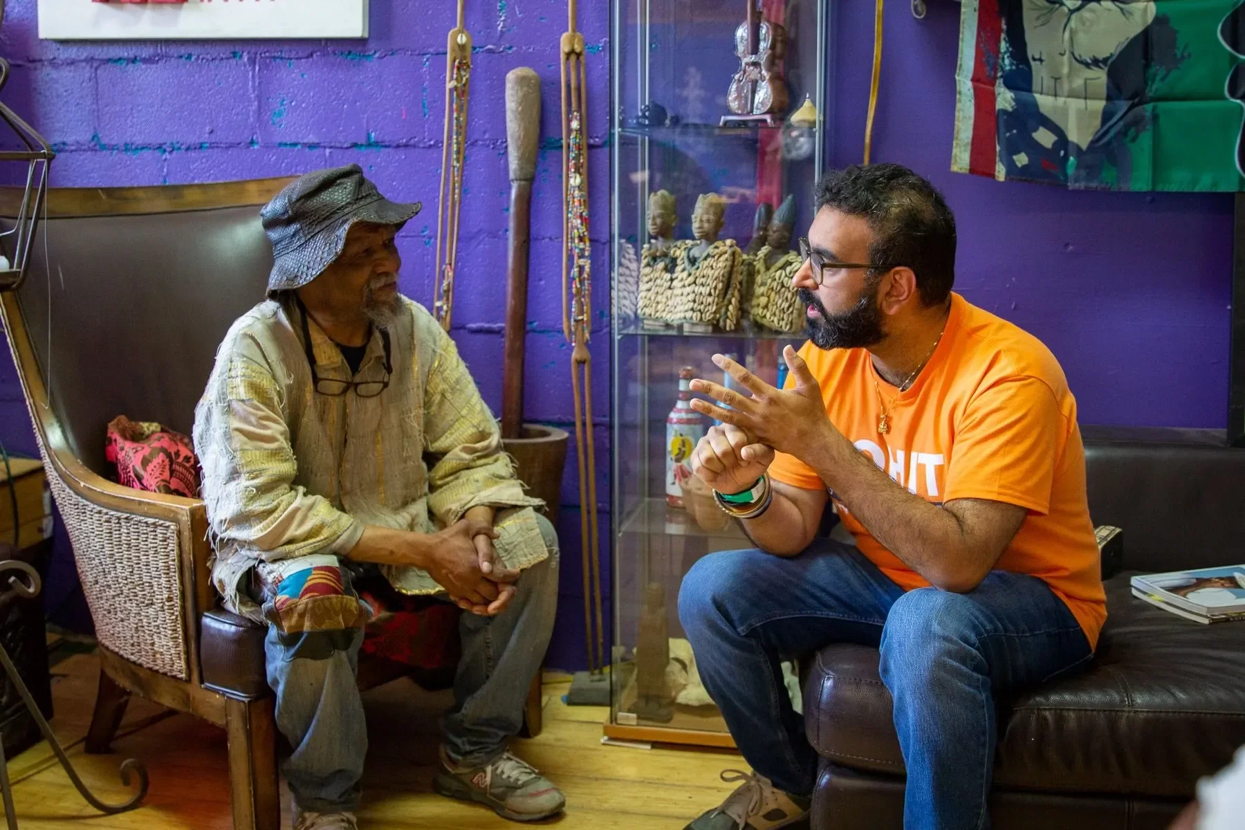 Rohit Malhotra and an elderly woman are sitting and conversing inside a colorful room decorated with African art and artifacts. They appear engaged in a friendly discussion.