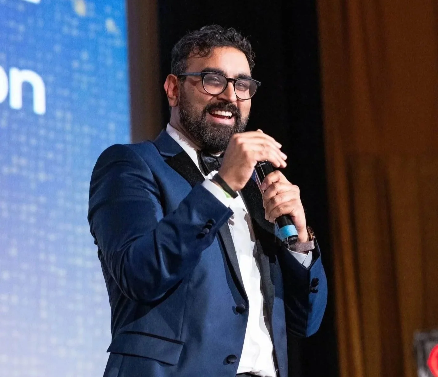Rohit Malhotra in a blue tuxedo with glasses and a beard speaking into a microphone at an event.