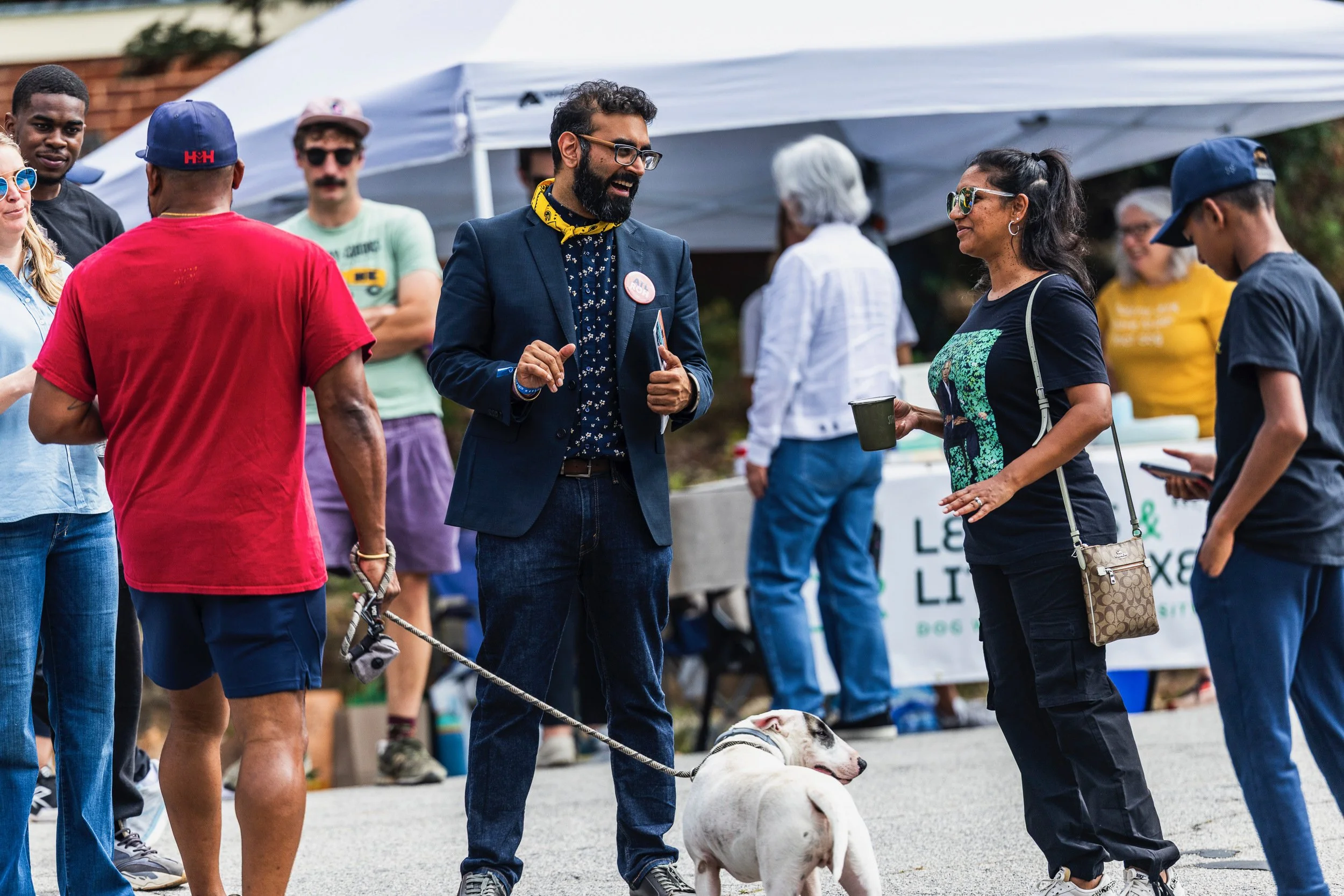 People gathered outdoors at a festival or event, some wearing sunglasses, with tents in the background. Rohit Malhotra in a suit with a yellow bandana around his neck is talking to a woman holding a mug, and a dog is on a leash nearby.