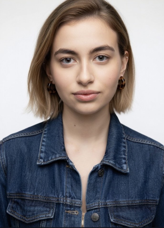 A young woman with short, light brown hair and blue eyes, wearing a denim jacket and earrings, standing against a plain white background.