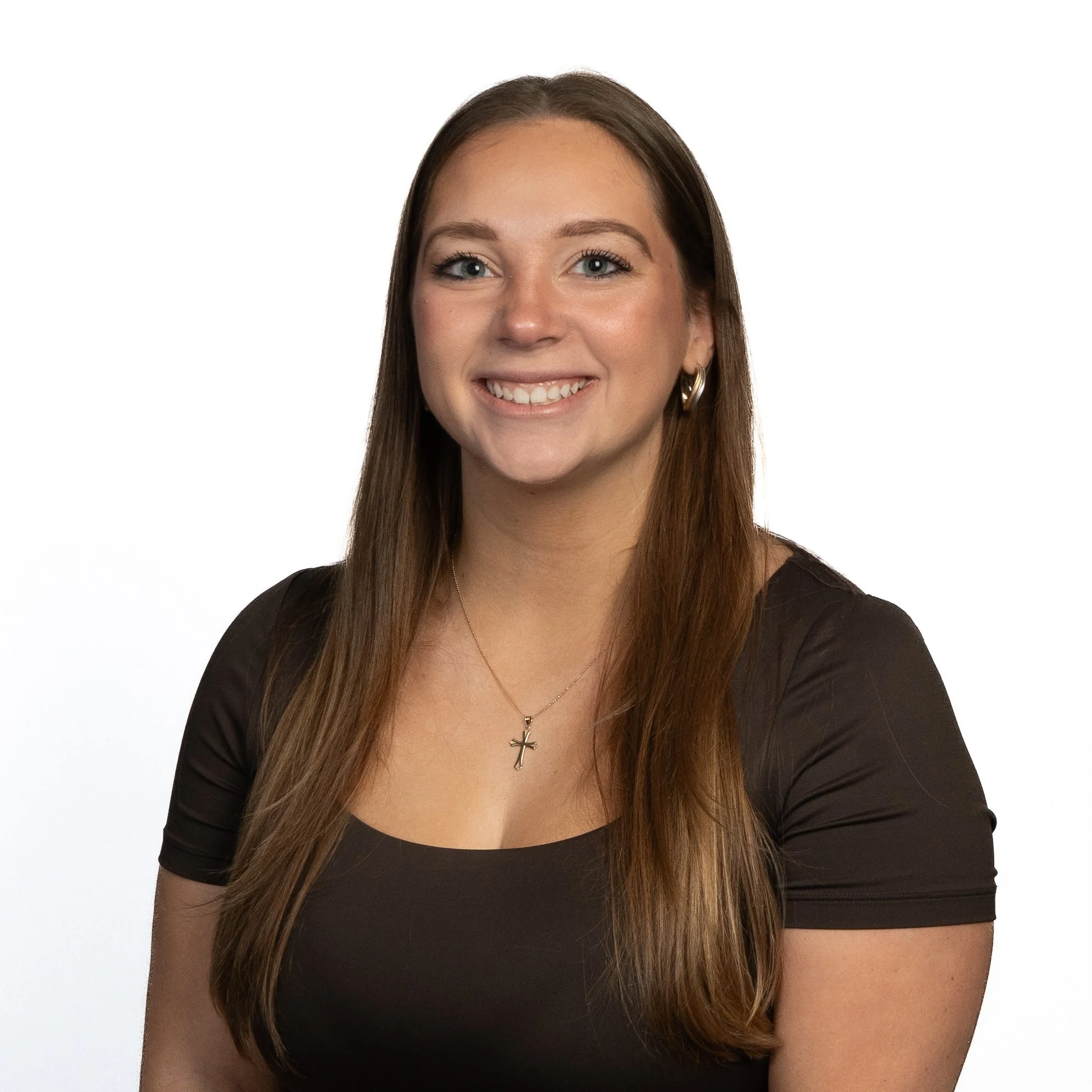 Smiling woman with long brown hair wearing a black top, gold hoop earrings, and a cross necklace, standing against a white background.