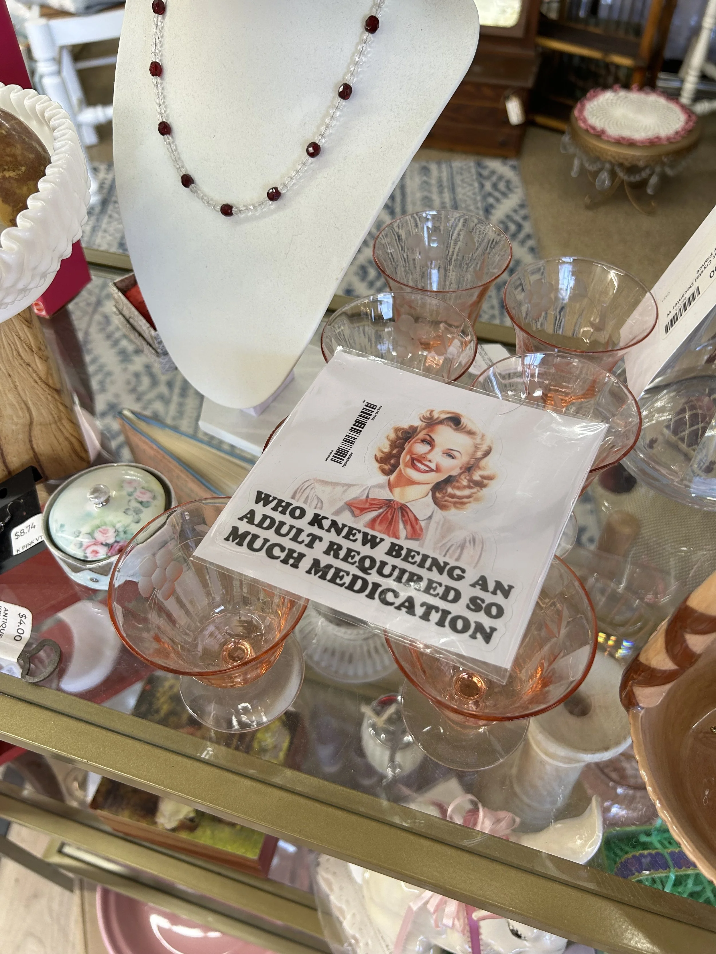 A display of glassware and jewelry in a thrift store. There are pink tinted glasses, a necklace with red beads, and a cardboard sign with a vintage woman and text about adult medical needs.