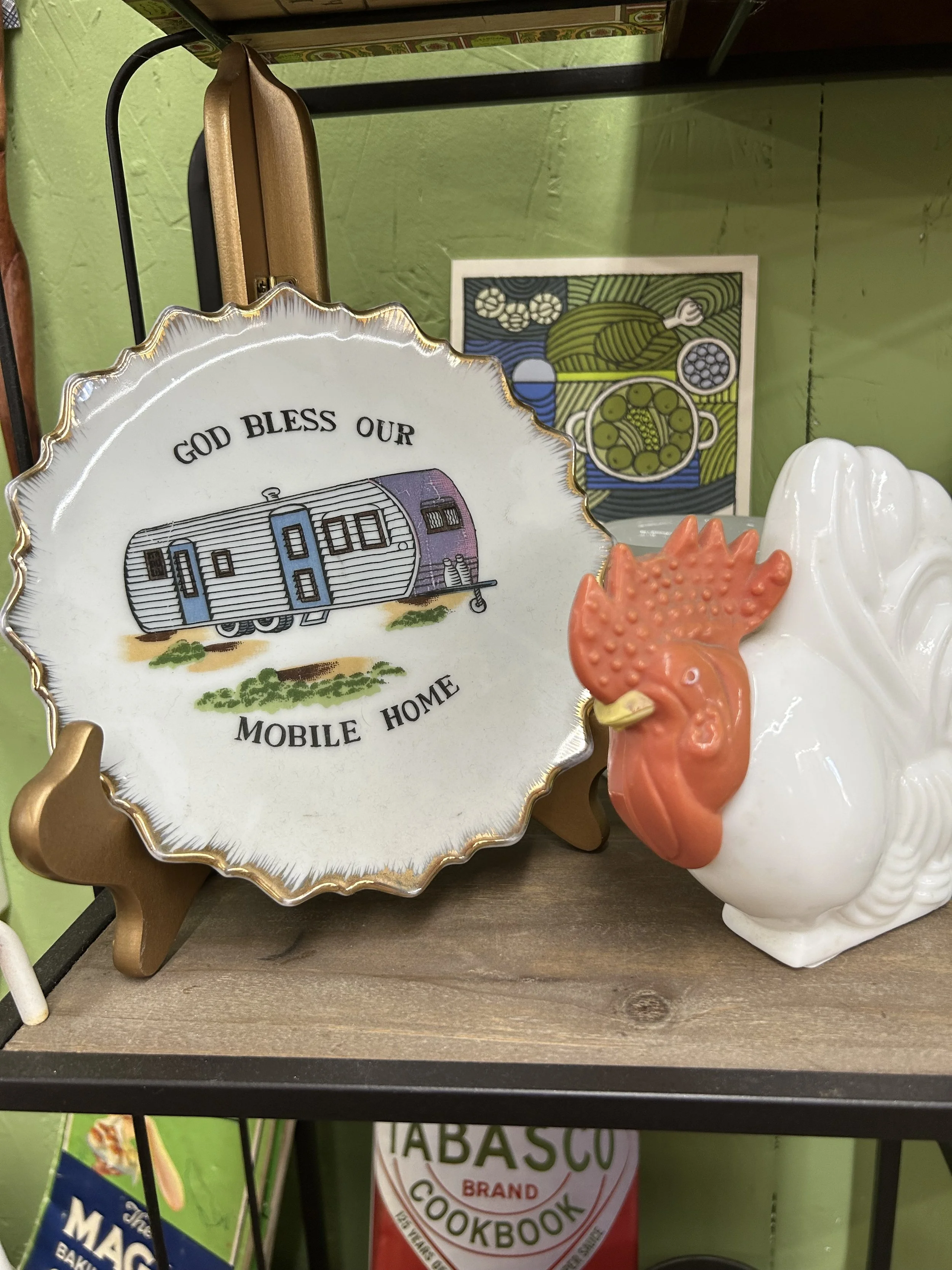Decorative ceramic plate with the phrase "God Bless Our Mobile Home" and an illustration of a travel trailer. A ceramic rooster figure is partially visible in front of the plate. In the background, there is a colorful artwork featuring a pot of green peas and other vegetables.