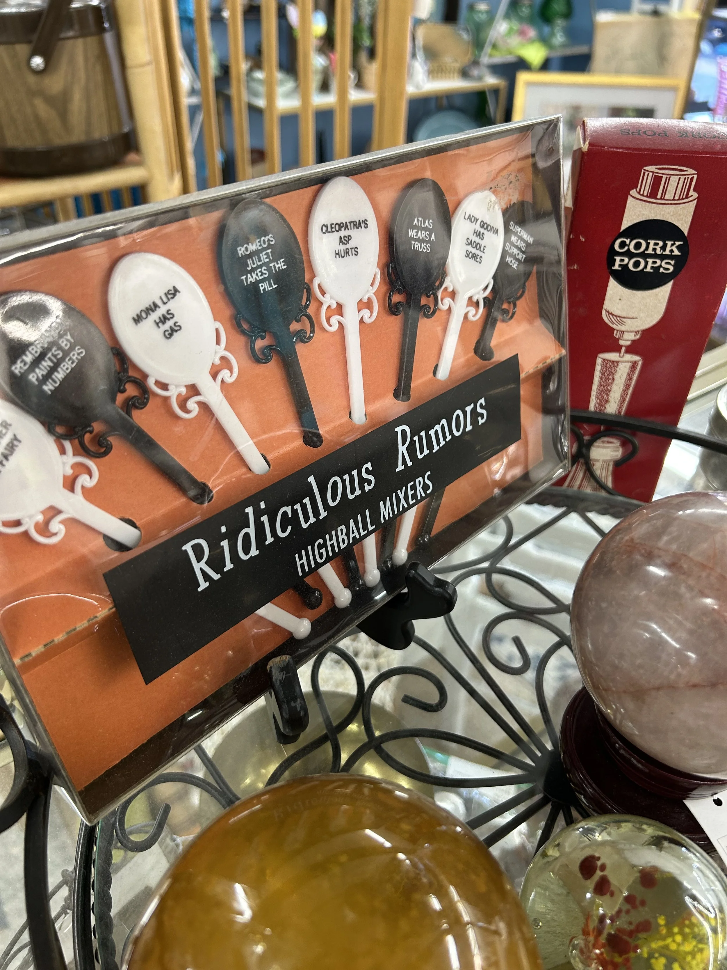 A display of novelty spatulas with humorous labels on a store shelf, with colorful jars in the foreground and a blurred background of store shelving.