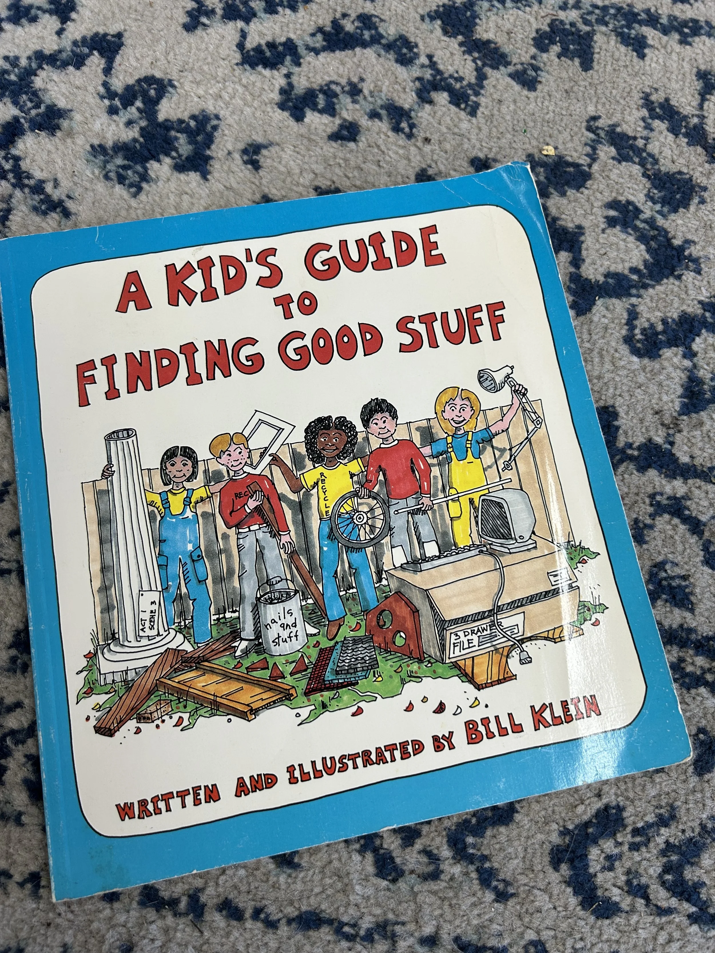 Book titled 'A Kid's Guide to Finding Good Stuff' resting on a carpeted floor with a blue, black, and beige pattern. The cover features cartoon children and a cluttered scene with various objects and debris.