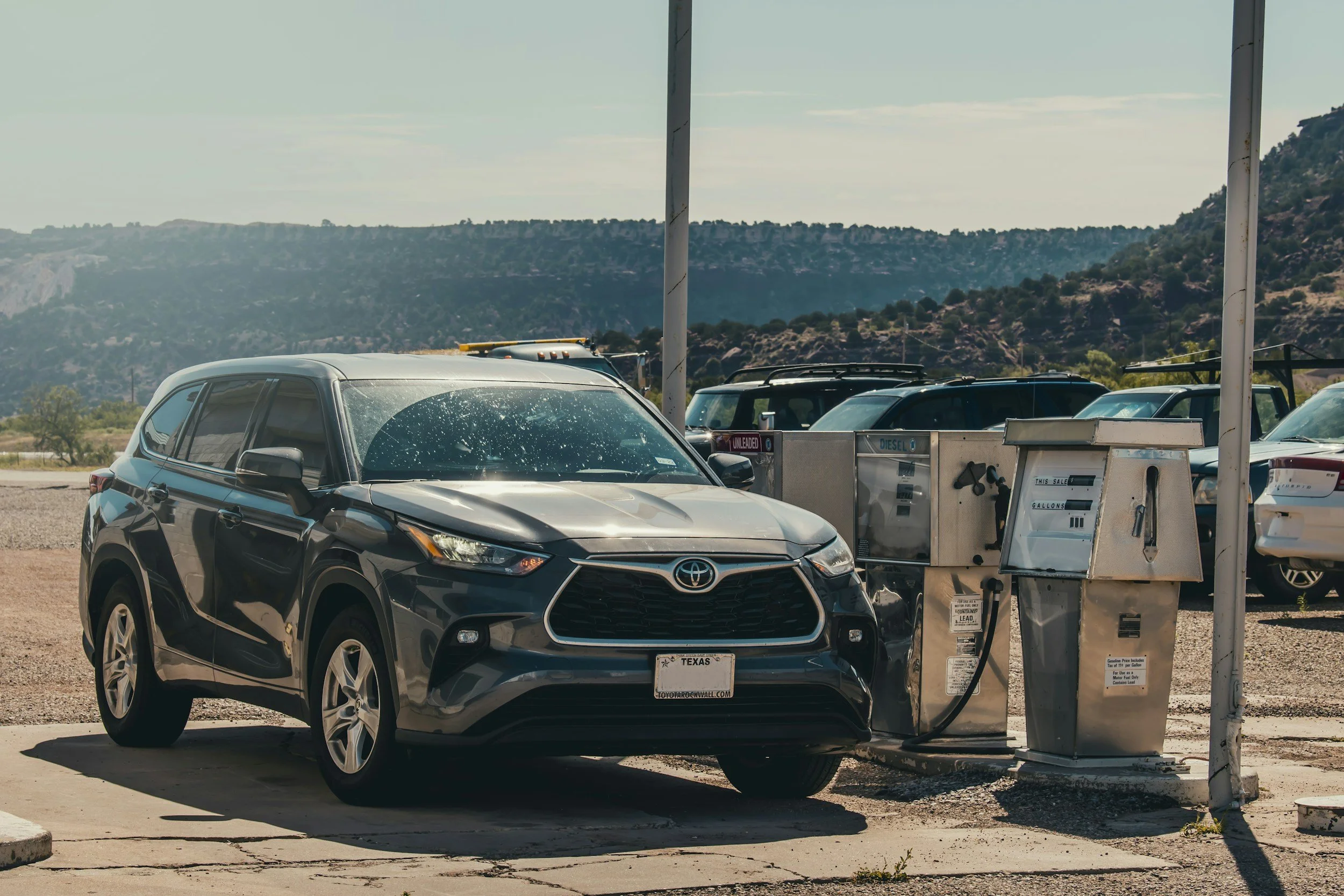A black Toyota SUV parked at a gas station in front of fuel pumps labeled for unleaded and diesel fuel, with a scenic mountain landscape in the background.