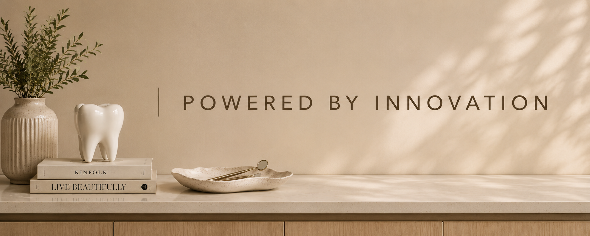 A still life photograph of a beige counter with a beige wall in the background displaying the text 'Powered by Innovation'. On the left, there is a beige textured vase with green foliage, a white decorative tooth sculpture, and two books titled 'Kinfolk' and 'Live Beautifully'. To the right of the books, there is a beige ceramic dish with a pair of silver tongs resting inside.