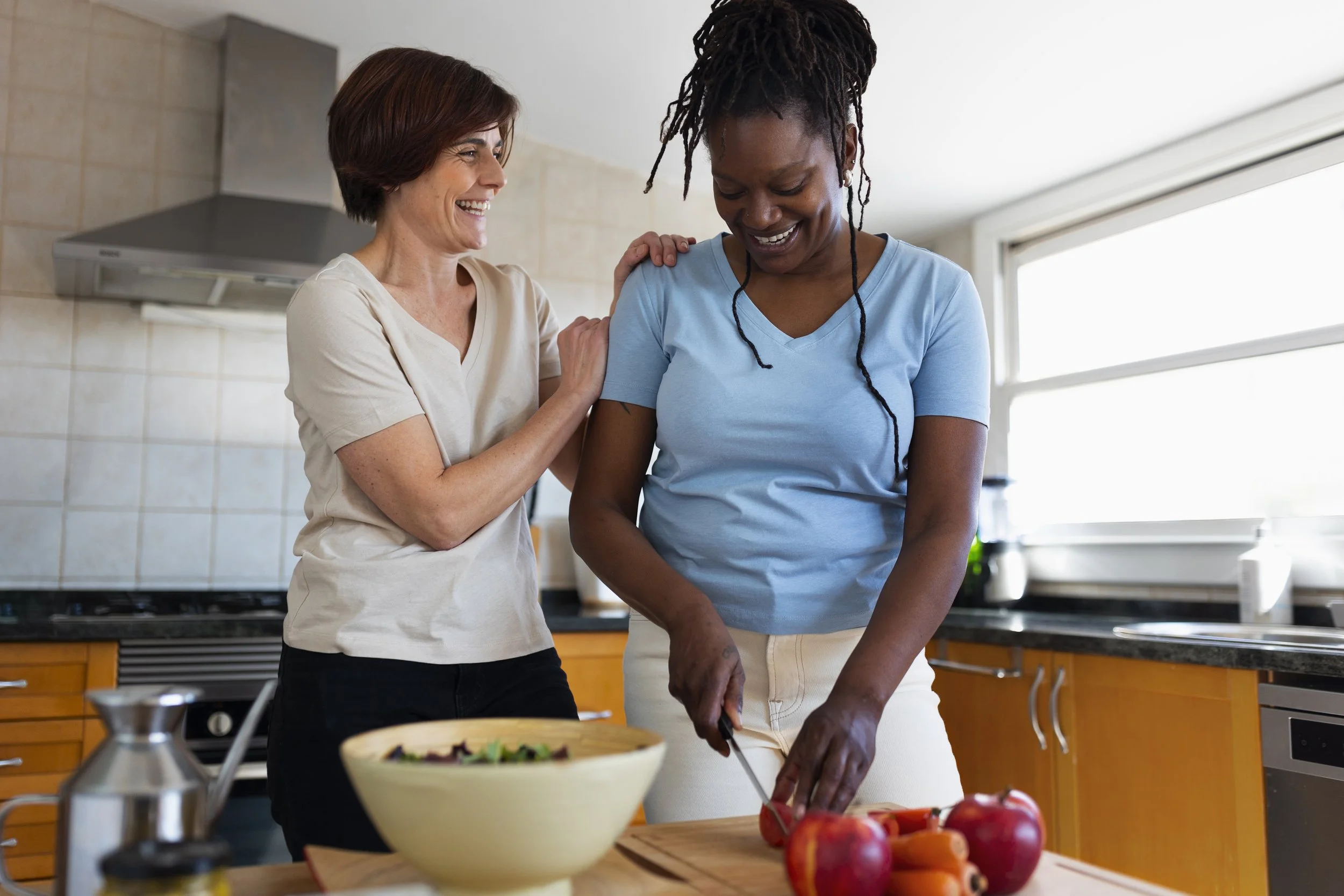 Two women, one older and one younger, are in a kitchen, smiling and preparing food together. The older woman is helping the younger woman cut tomatoes with a knife on a cutting board. There is a salad in a bowl, apples, and carrots on the countertop.
